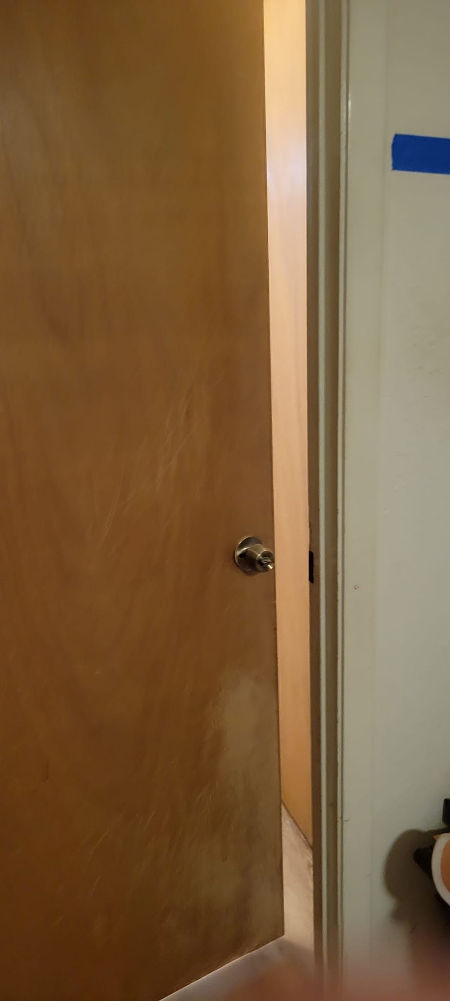 Ajar wooden door with a doorknob, revealing a brightly lit doorway. A wall with blue tape is visible to the right.