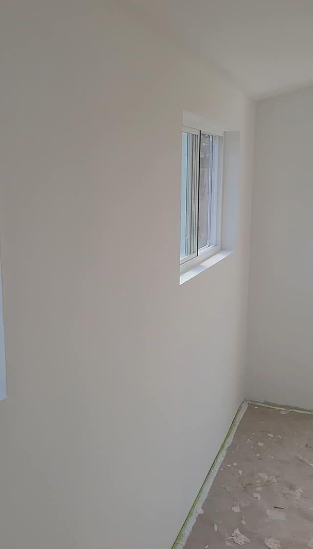 Interior view of a room with white walls, a window with blinds, and a concrete floor. Green tape lines the baseboards.
