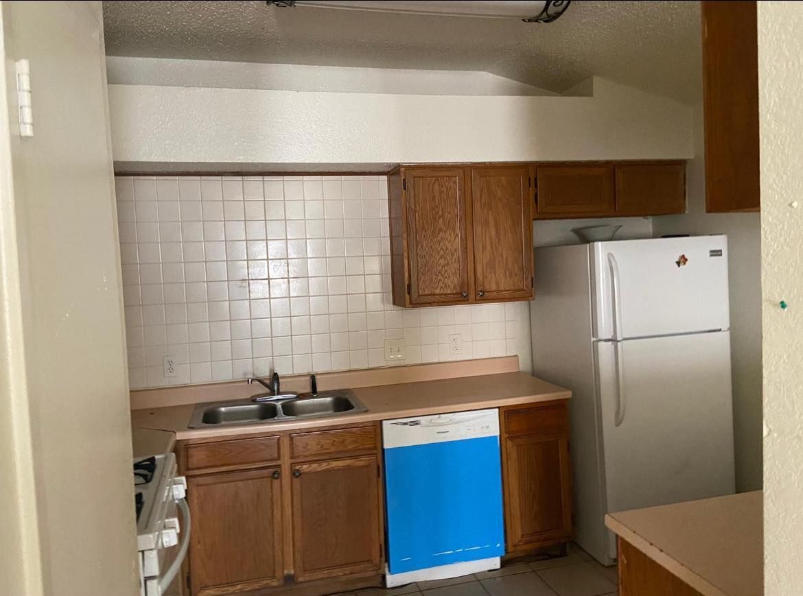 A small kitchen with wooden cabinets, a white refrigerator, a blue dishwasher, and a stainless steel sink. 