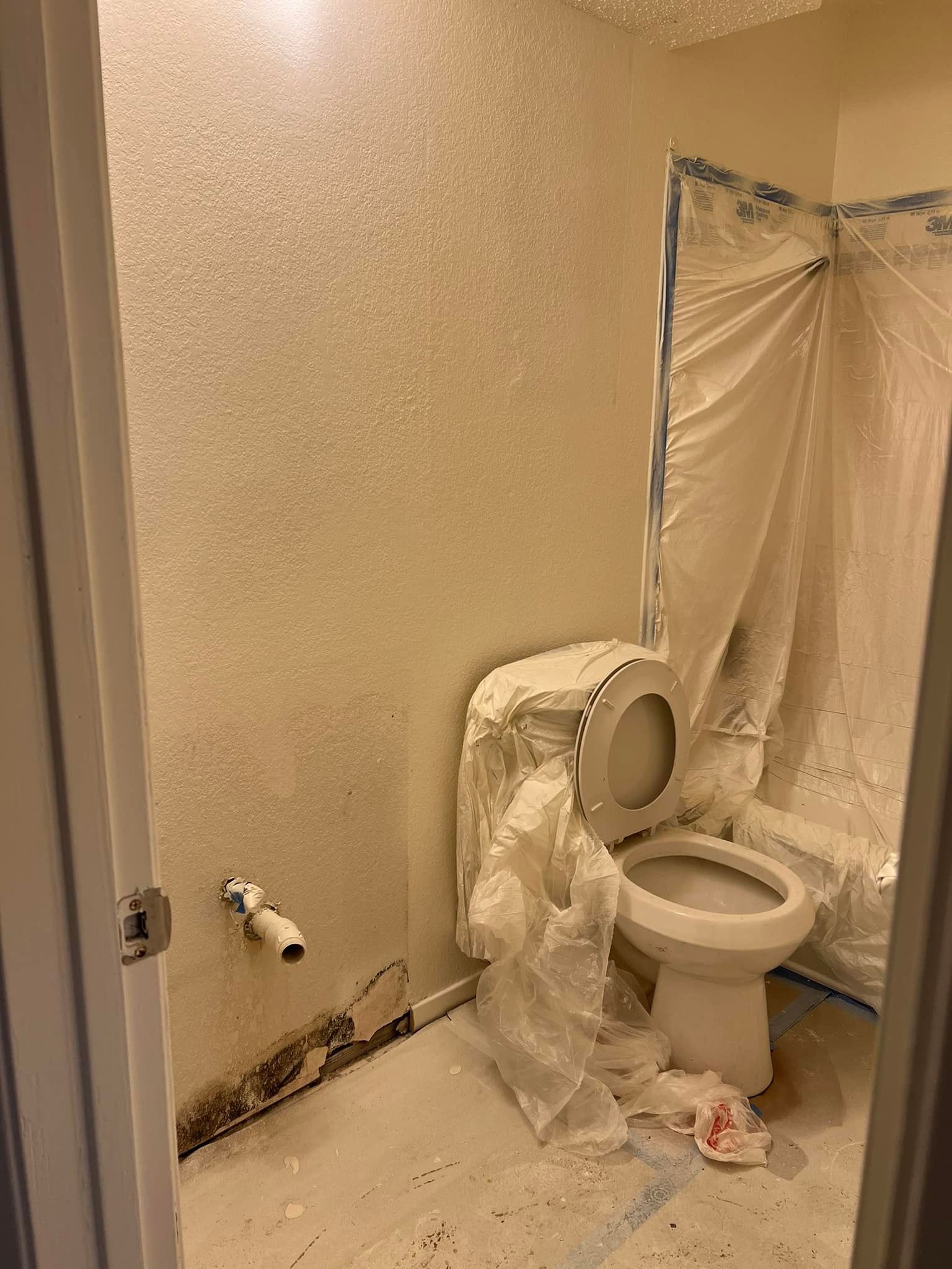 A small bathroom with white textured walls. Toilet and shower are covered in plastic sheeting. The floor has debris.