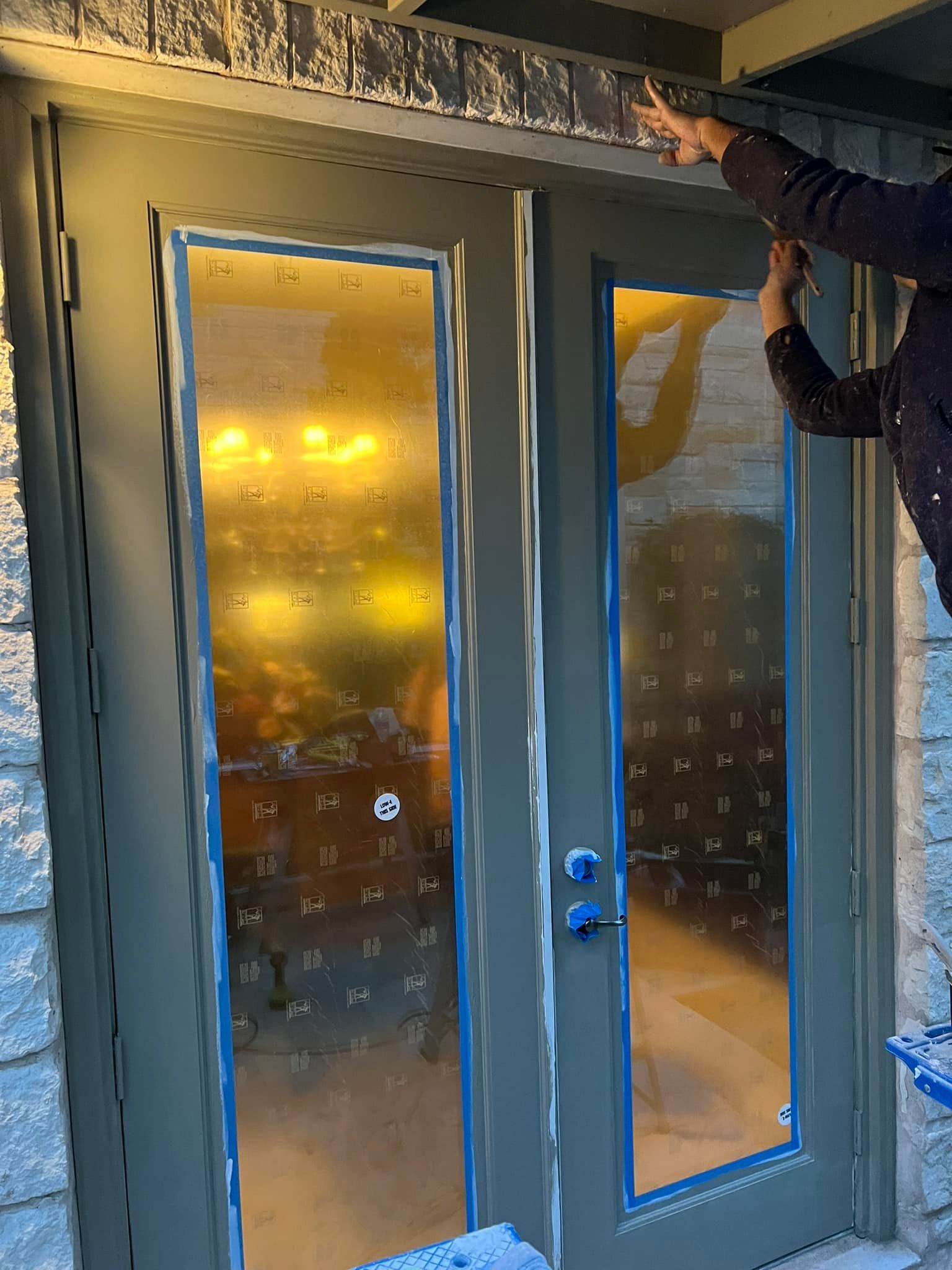 Person taping trim above a grey double door with glass panes. The door frame and surrounding siding are also grey.