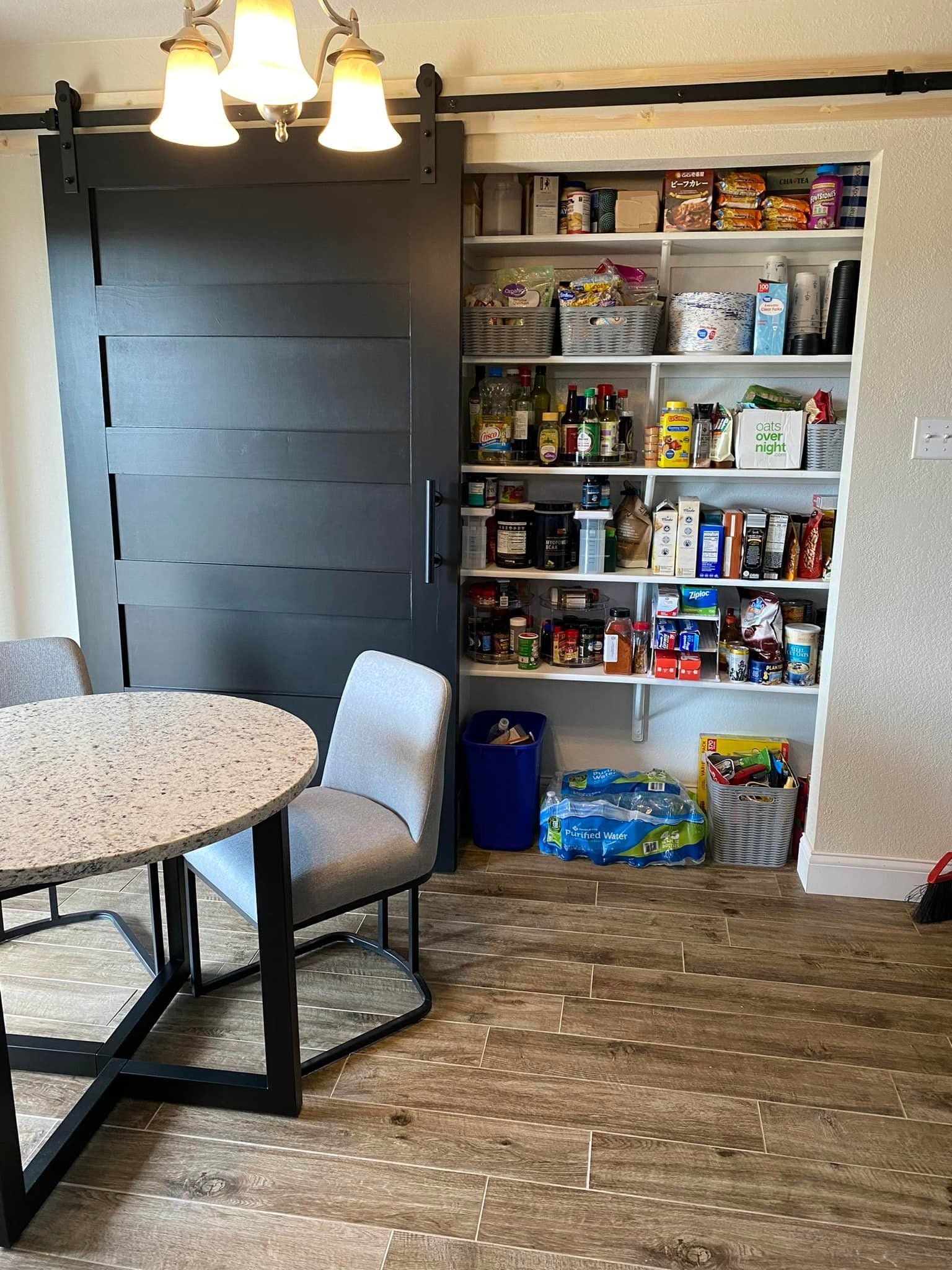 A dining area with a round table and chairs next to a pantry with a sliding barn door. The pantry is filled with food items.