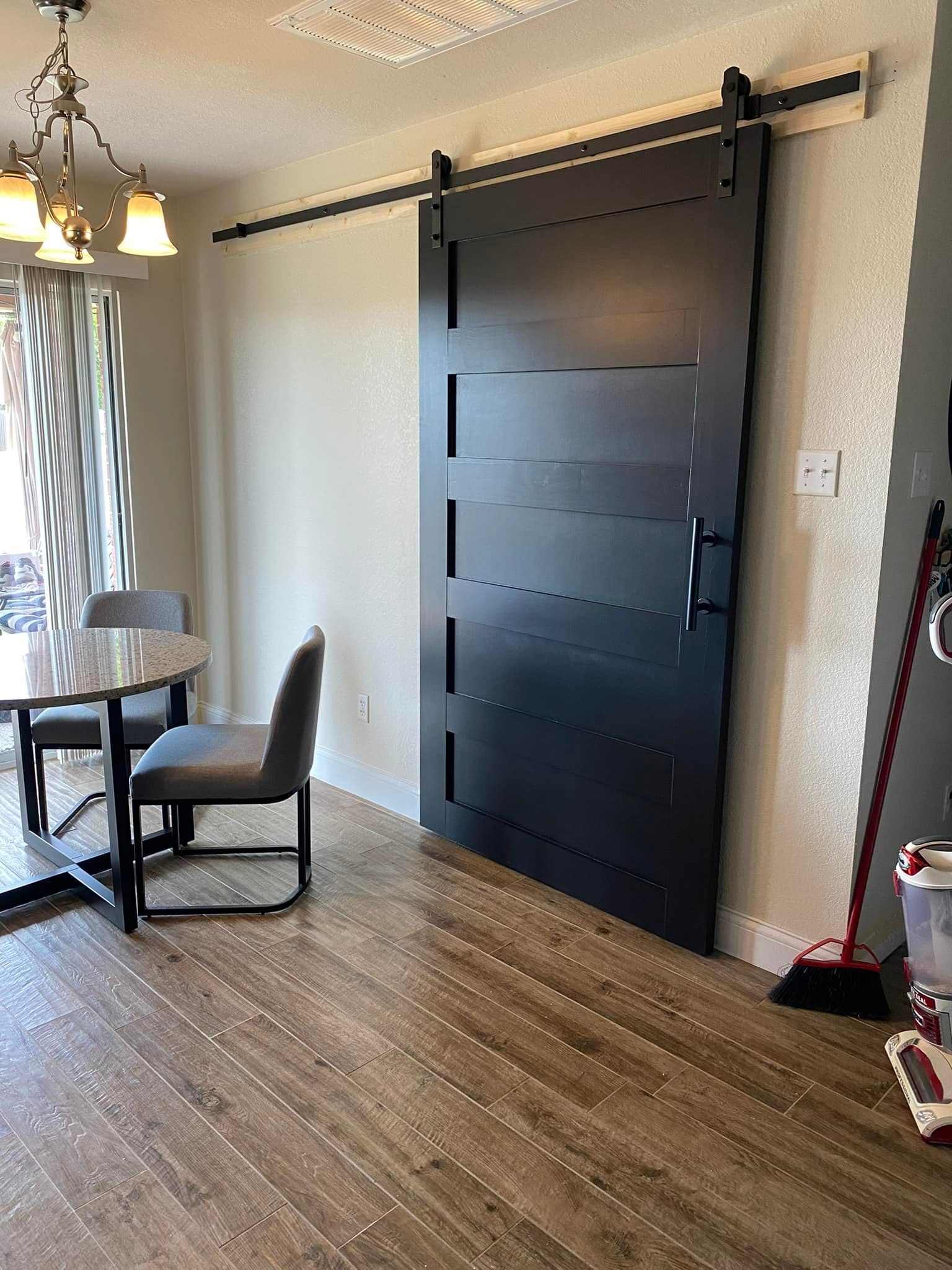 A black, horizontal-paneled barn door in a dining area, with a table and chairs to the left, and a broom on the right.