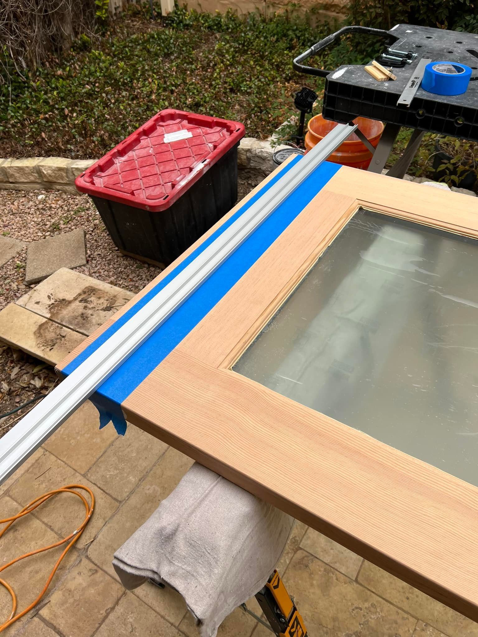 A person is working on a wooden door frame with a metal guide, tape, and tools outdoors. A red bin is nearby.