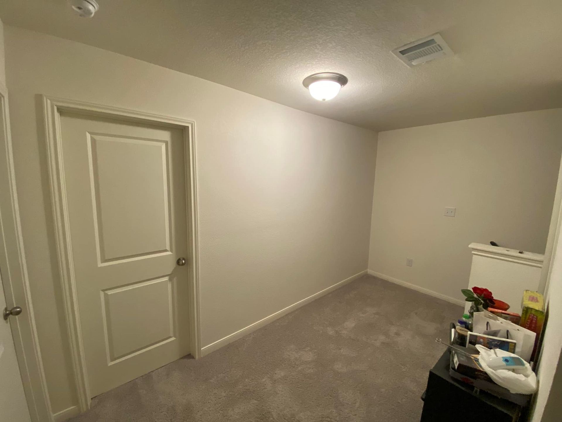 Small room with carpet, a closed door, and a ceiling light. There is a black dresser with items on it.