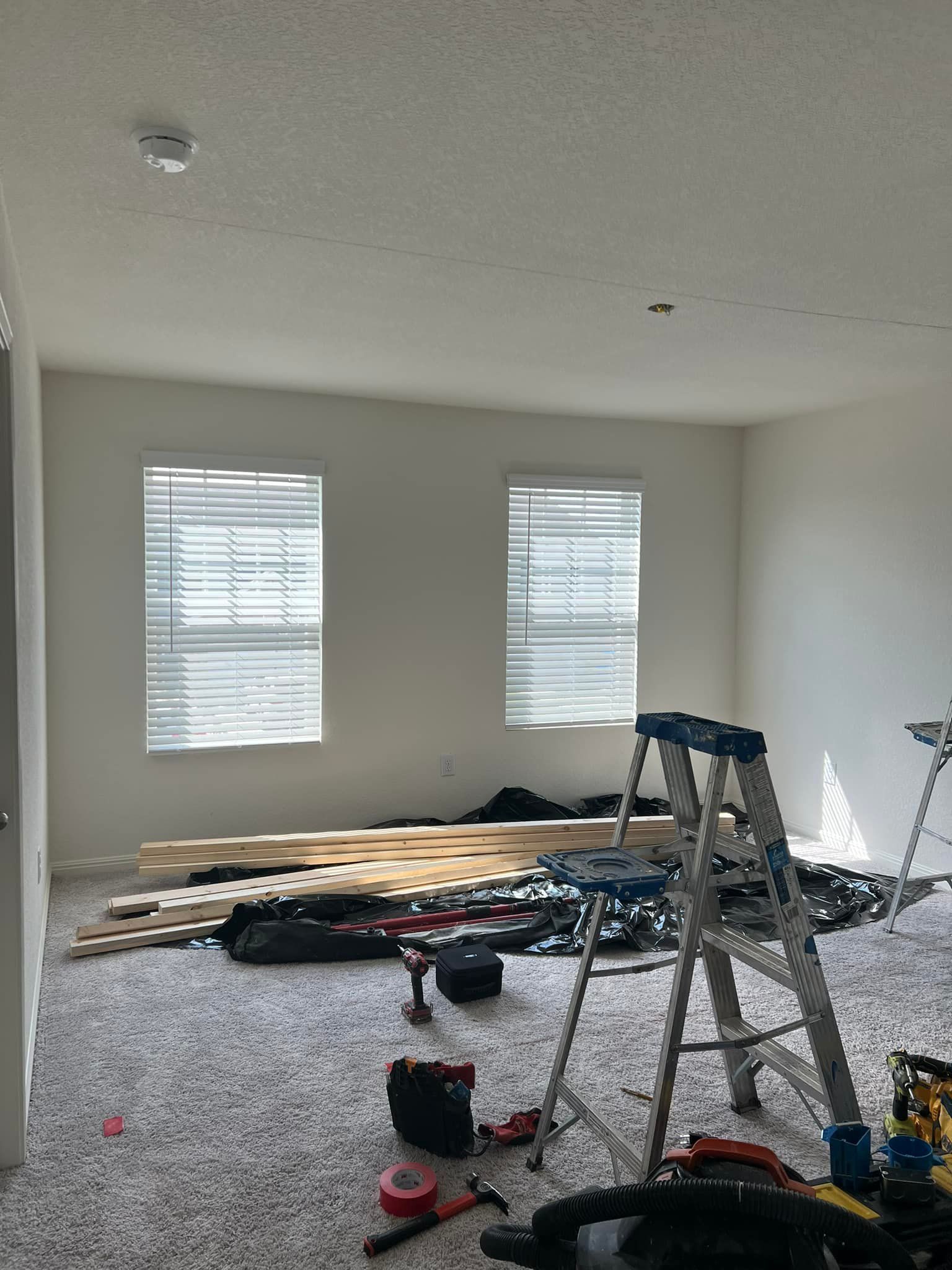 A room under construction with two windows, a ladder, tools, and wood planks on the floor.