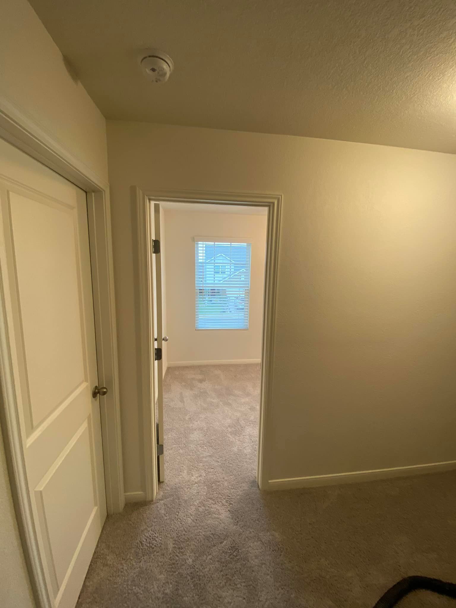 An open doorway leads into a room with a window. A closed door is on the left, all in a room with tan carpet and white walls.
