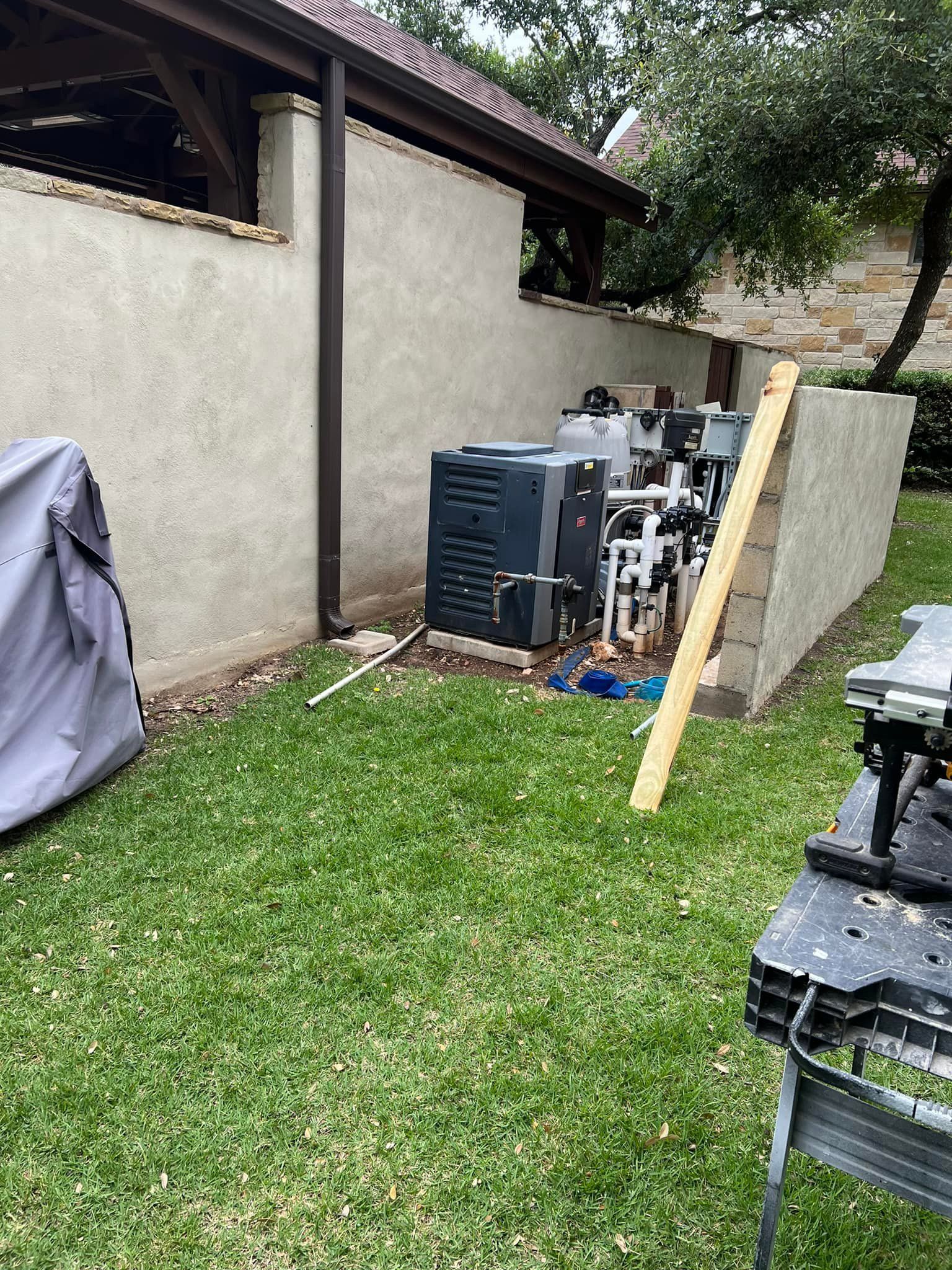Backyard view of pool equipment, including a heater and filter, partially obscured by a stucco wall & covered by a gray tarp.