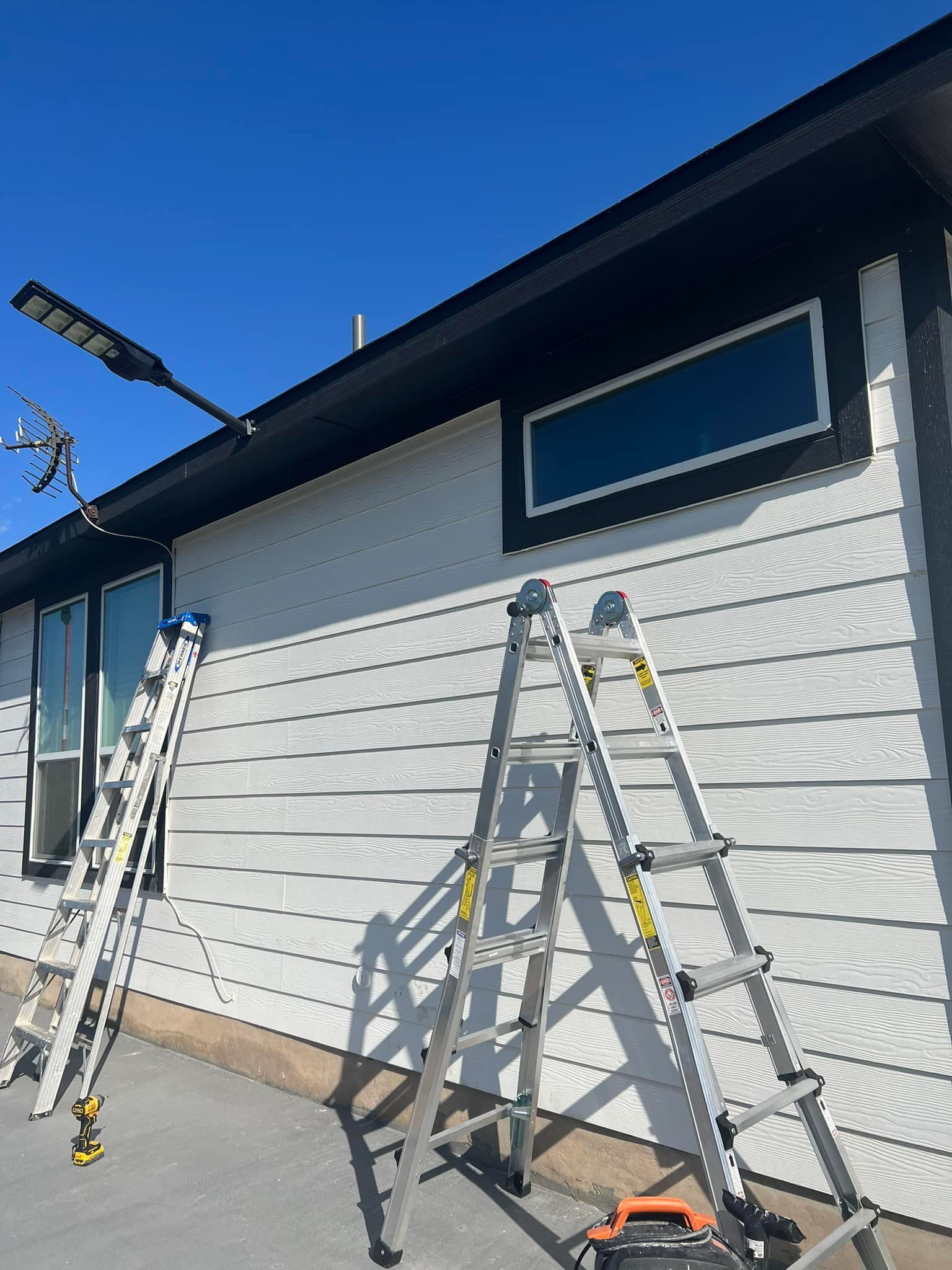Two ladders lean against a white-sided building with a black trim and a rectangular window with tinted glass.