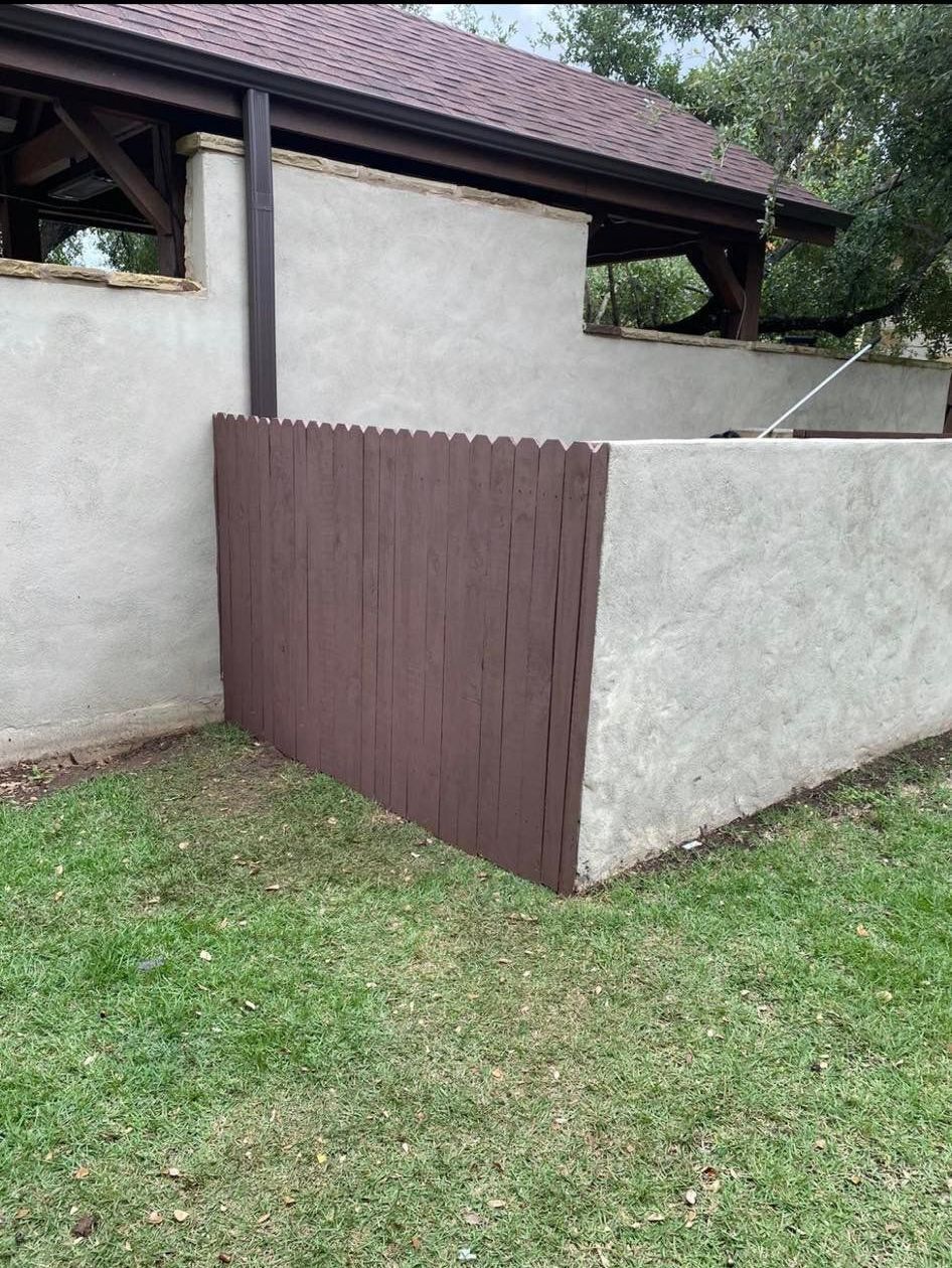 Brown fence section attached to a stucco wall, partially shaded by a brown-roofed structure; green grass in front.