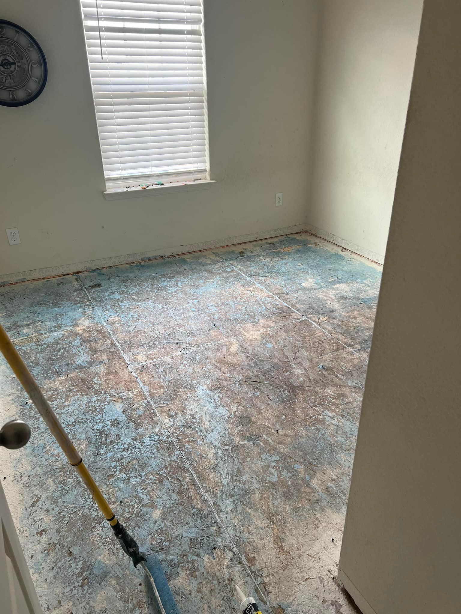 Room with a worn, patterned floor. A window with blinds lets in light. A broom is in the foreground.