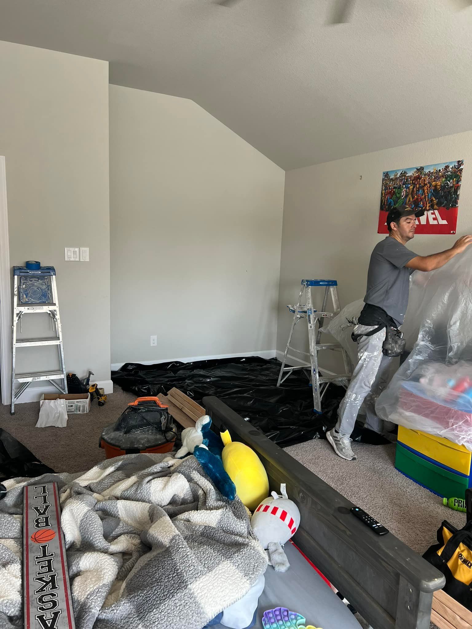 Man in a messy room, standing near a covered object, possibly moving items. A ladder, tarp, and bed are also in the room.