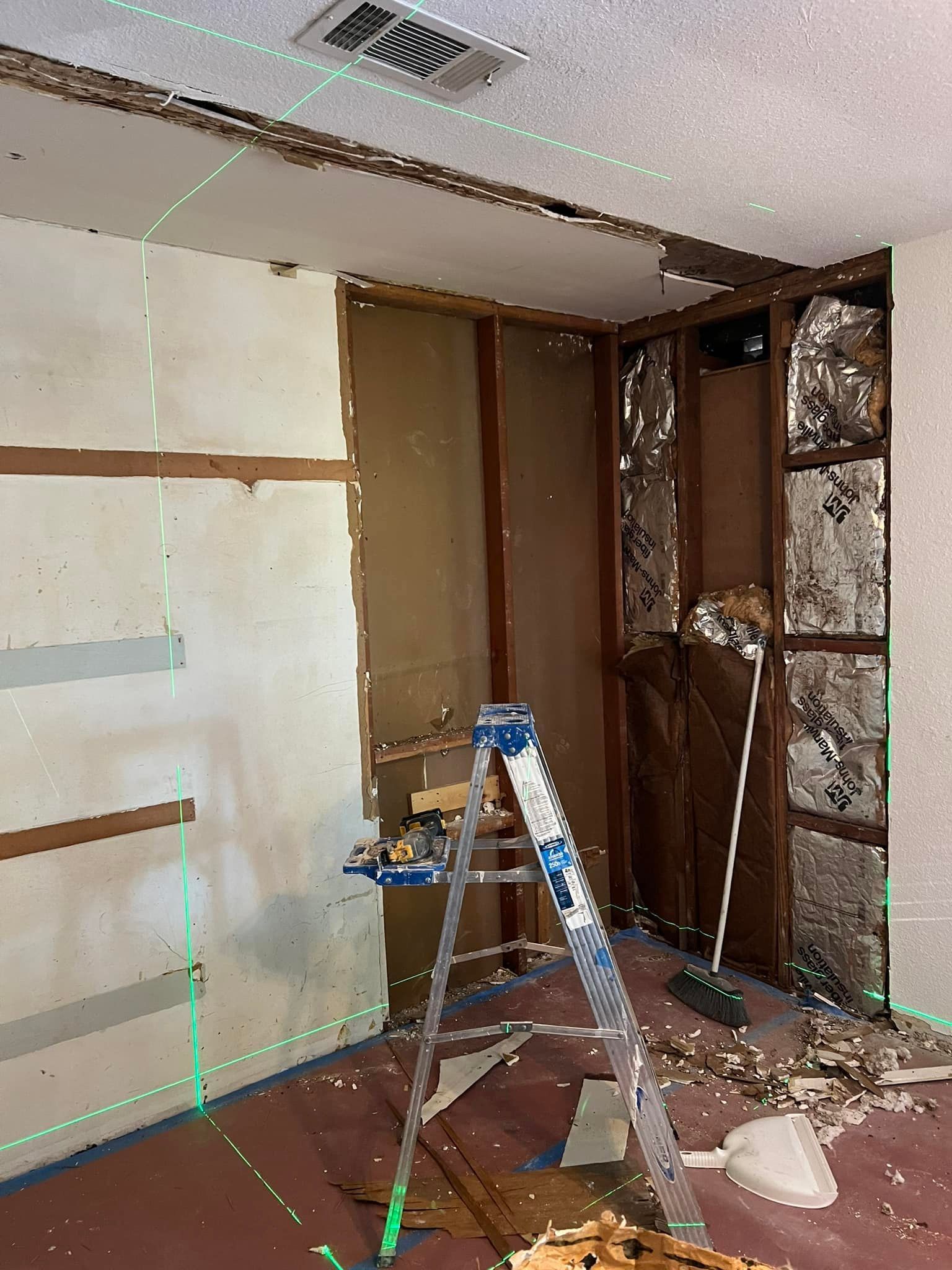 Interior demolition with exposed framing, insulation, and a ladder. A green laser level guides the work.