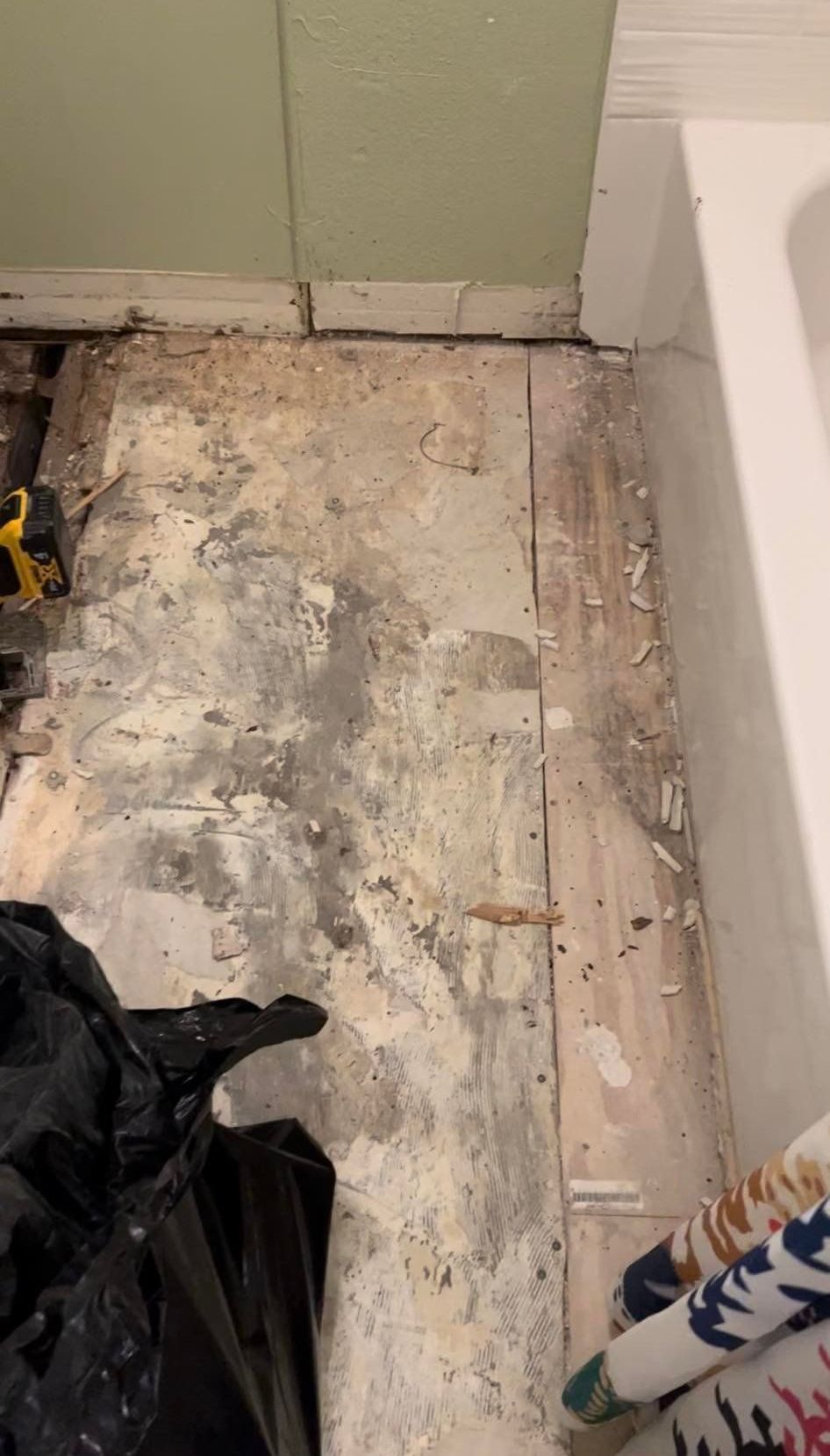 Damaged wooden floorboards in a room, with debris, a black trash bag, and tools visible.