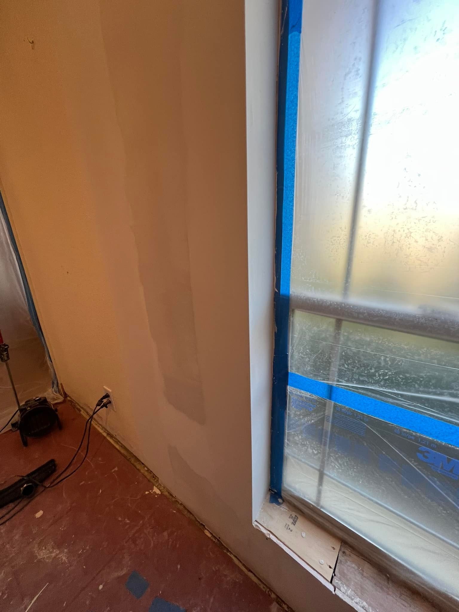 Interior view of a room with a window covered in blue tape. The wall is painted beige and the floor is red.