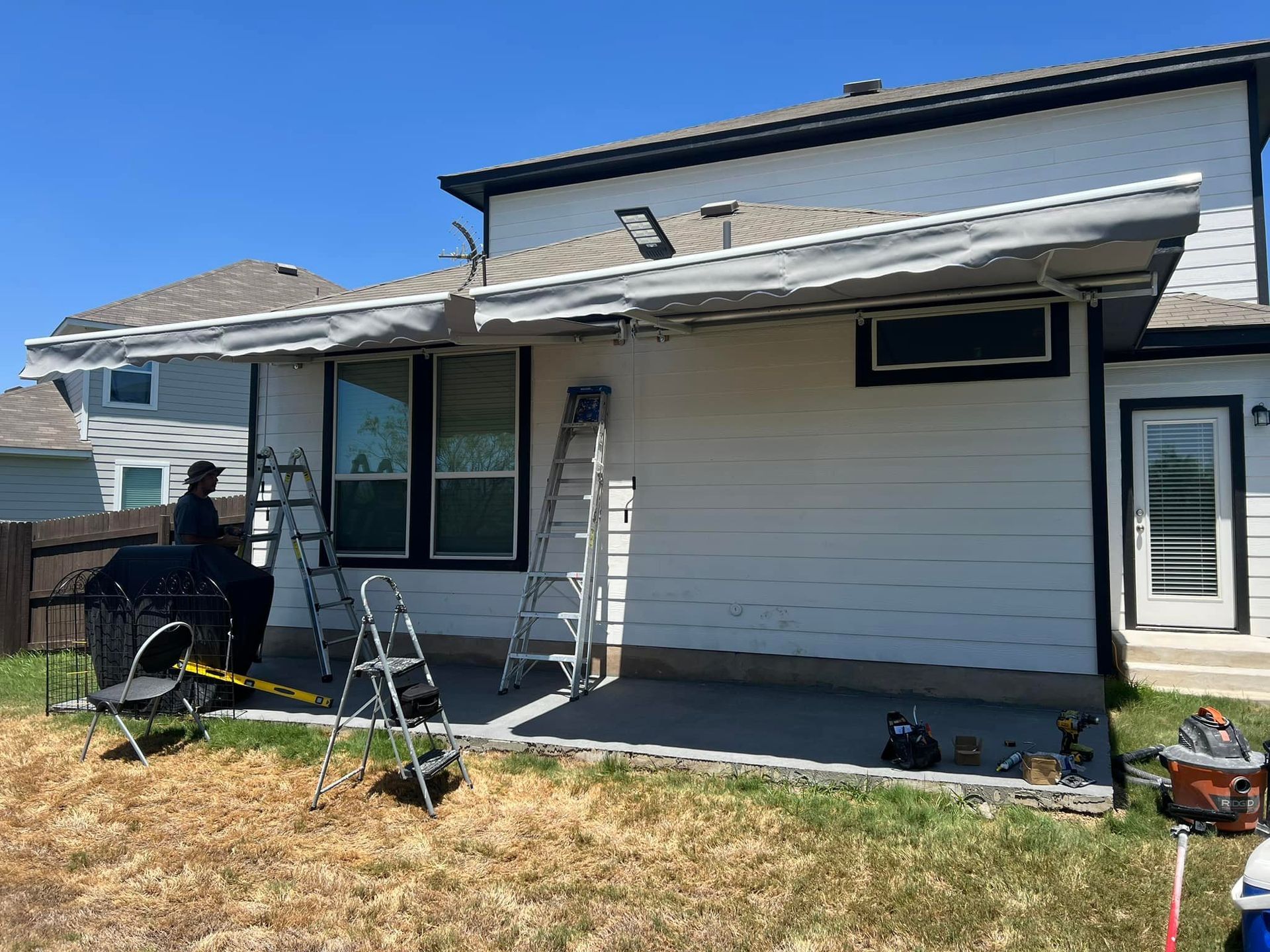 A person working on a white house with an awning installed. A ladder and tools are present on the patio.