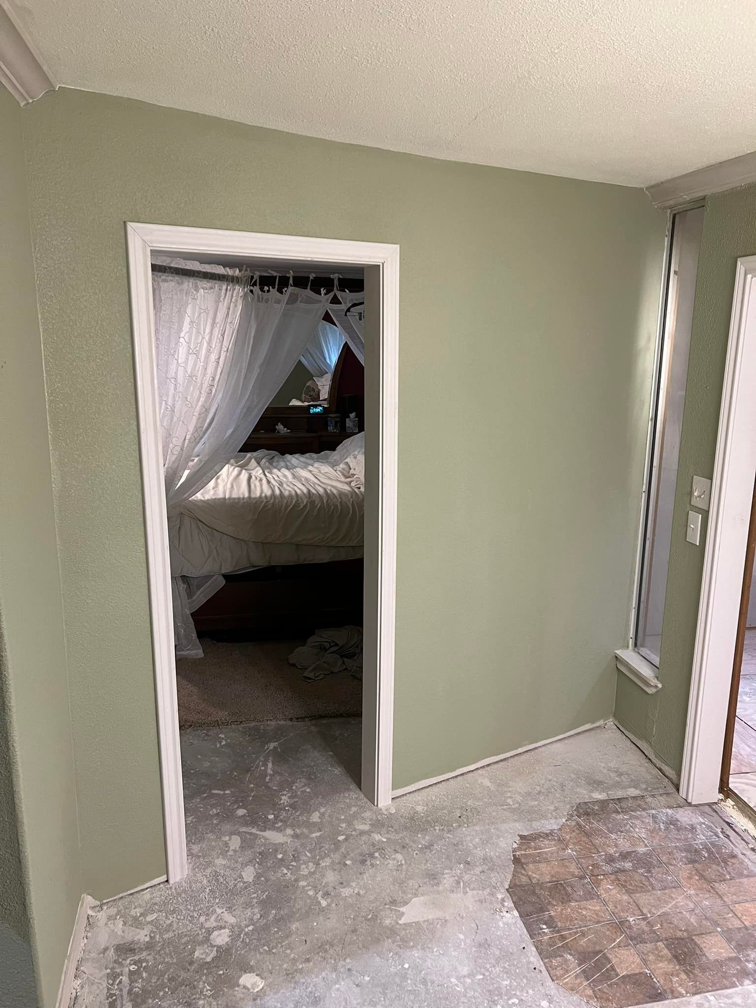 A doorway with white trim leads to a bedroom with a bed and curtains. The walls are light green with debris on the floor.