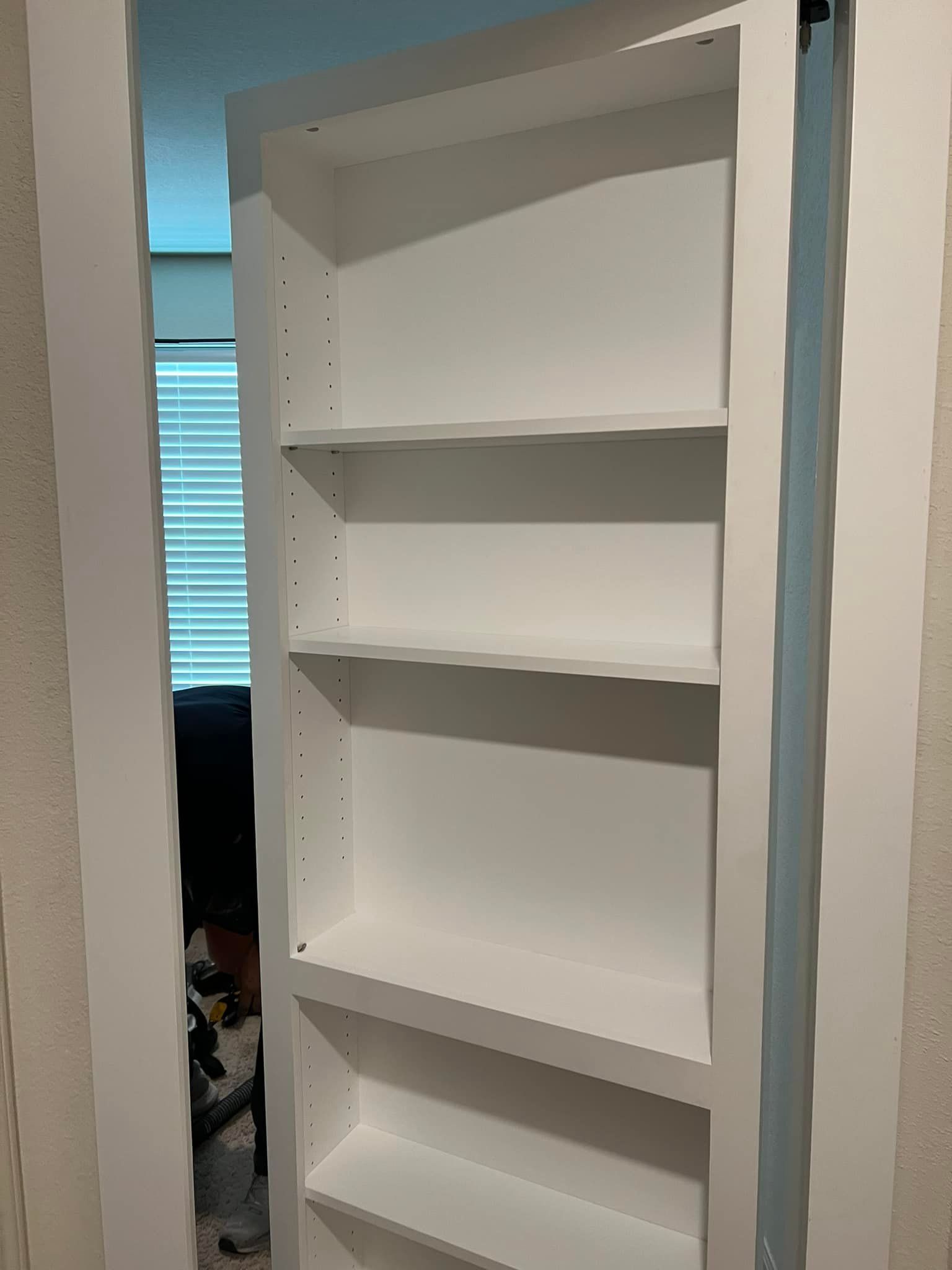White bookshelf built into a wall, with four shelves and an opening to a room with window blinds.