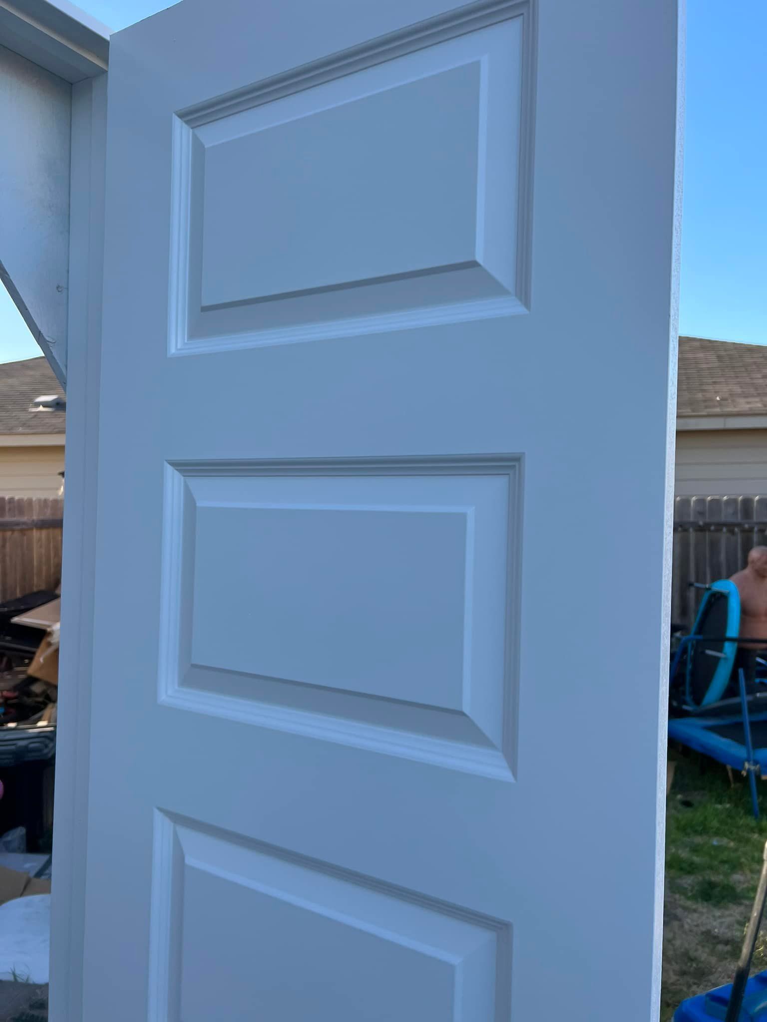 White three-panel door outdoors against a fence and clear sky. The door is leaning against a structure.
