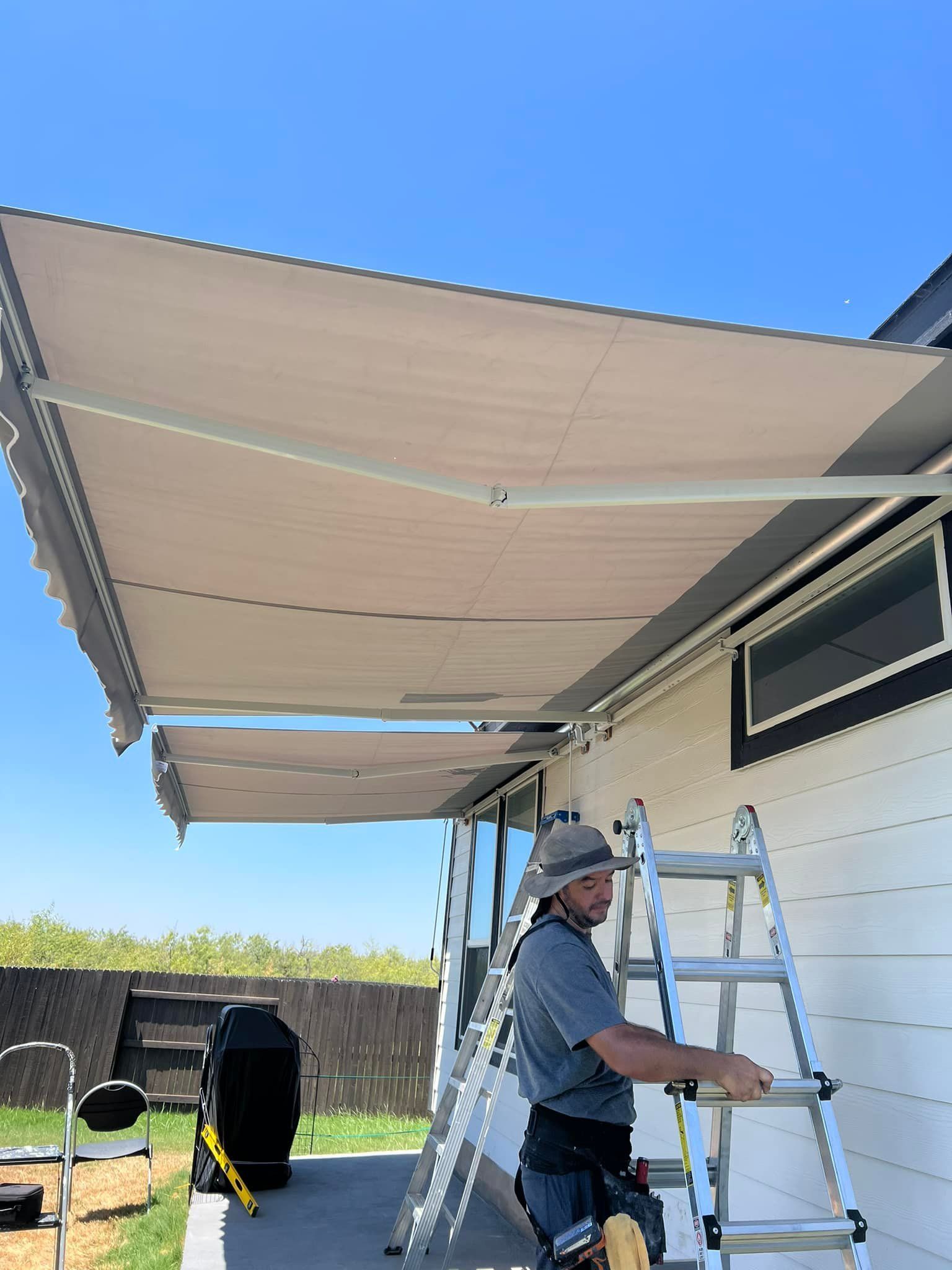 Man on a ladder installing a retractable awning on a white building under a clear blue sky.