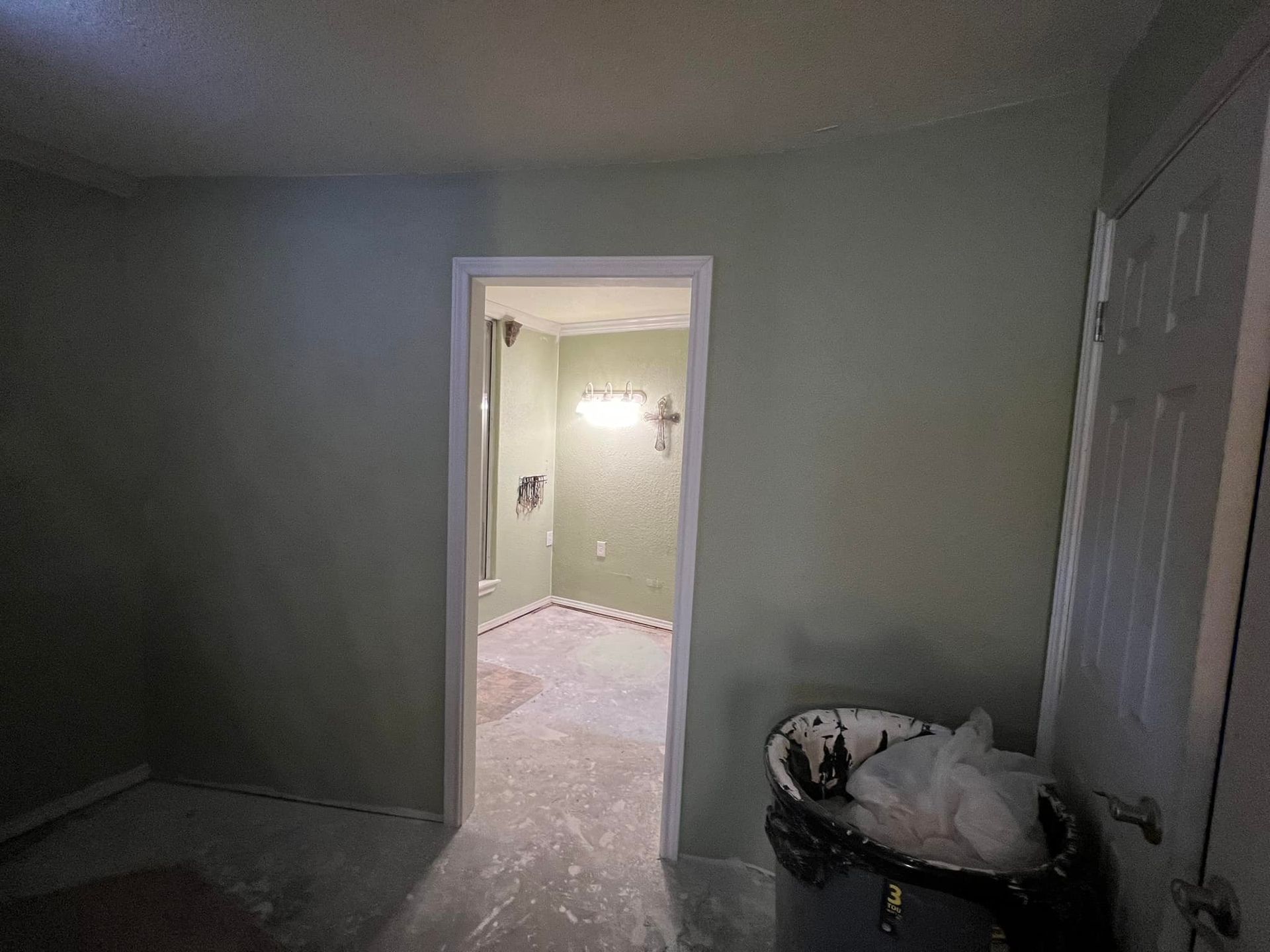 A small bathroom with light green walls and doorway, trash can visible in foreground.