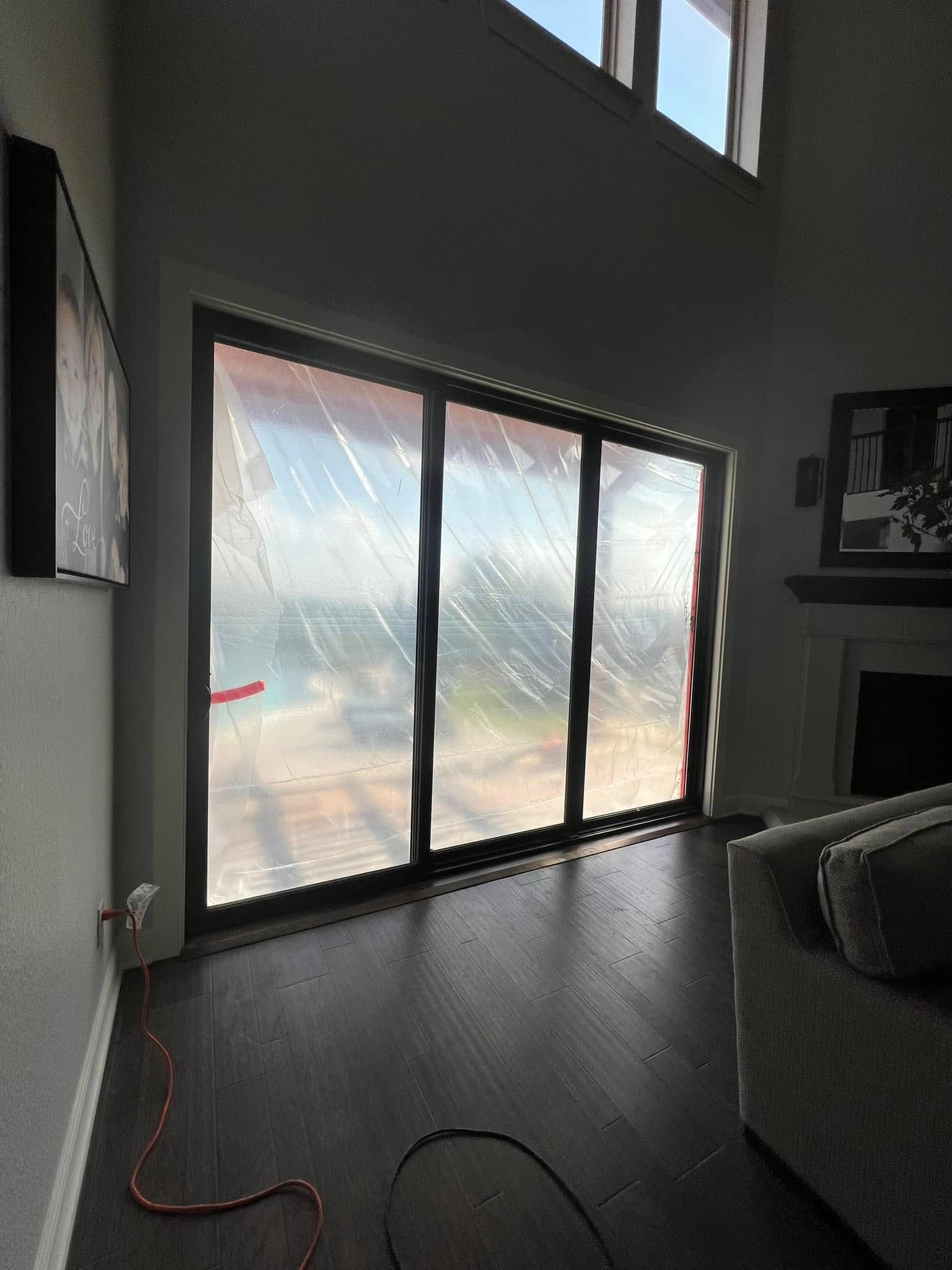A large, sliding glass door covered in protective plastic film, inside a living room with dark wood floors.
