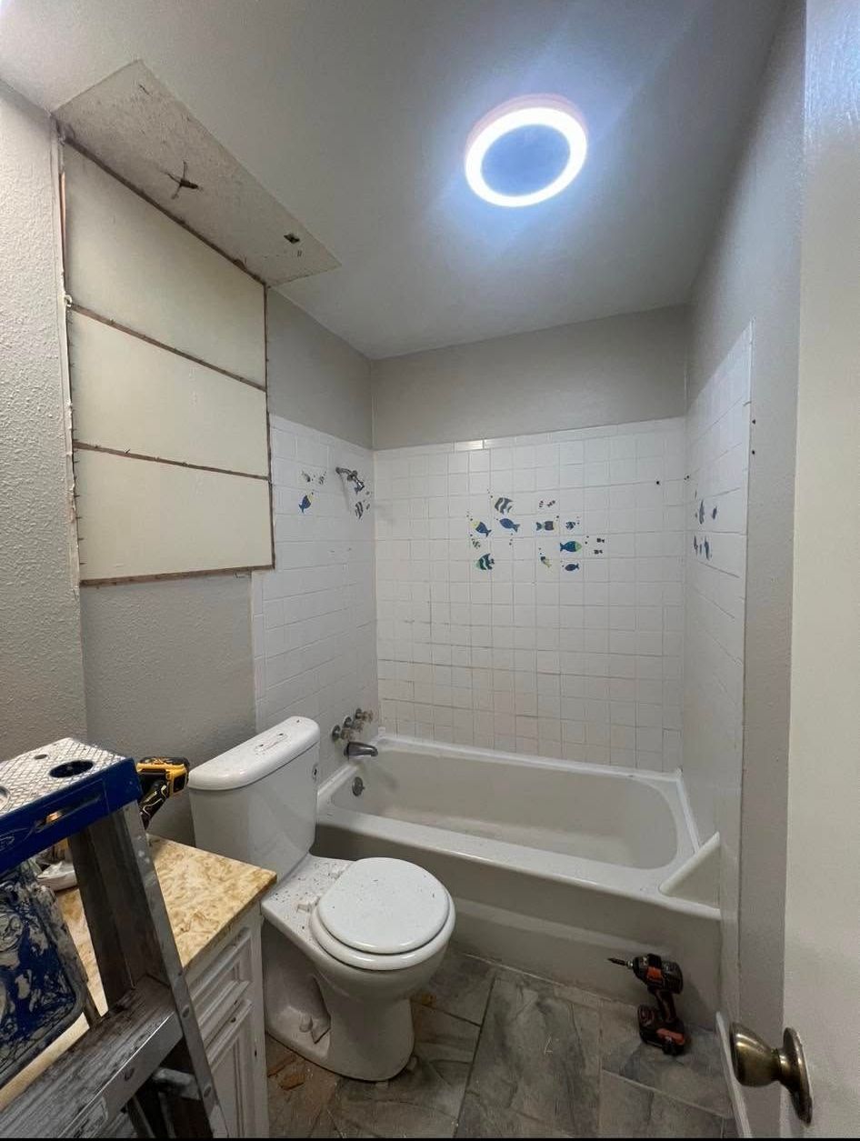 Bathroom with white tub, toilet, and tiled wall. A ladder and tools are in the foreground.