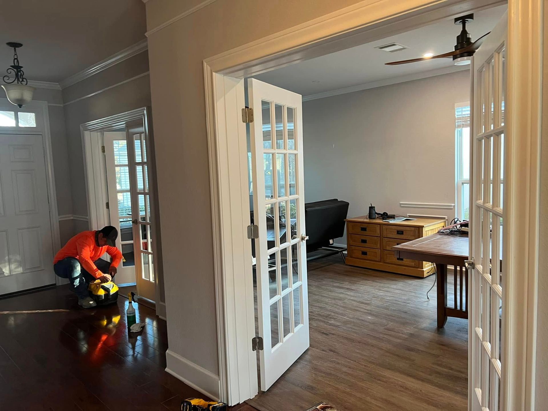 Man working on flooring near French doors in a home. The doors lead to a living room with furniture and a ceiling fan.