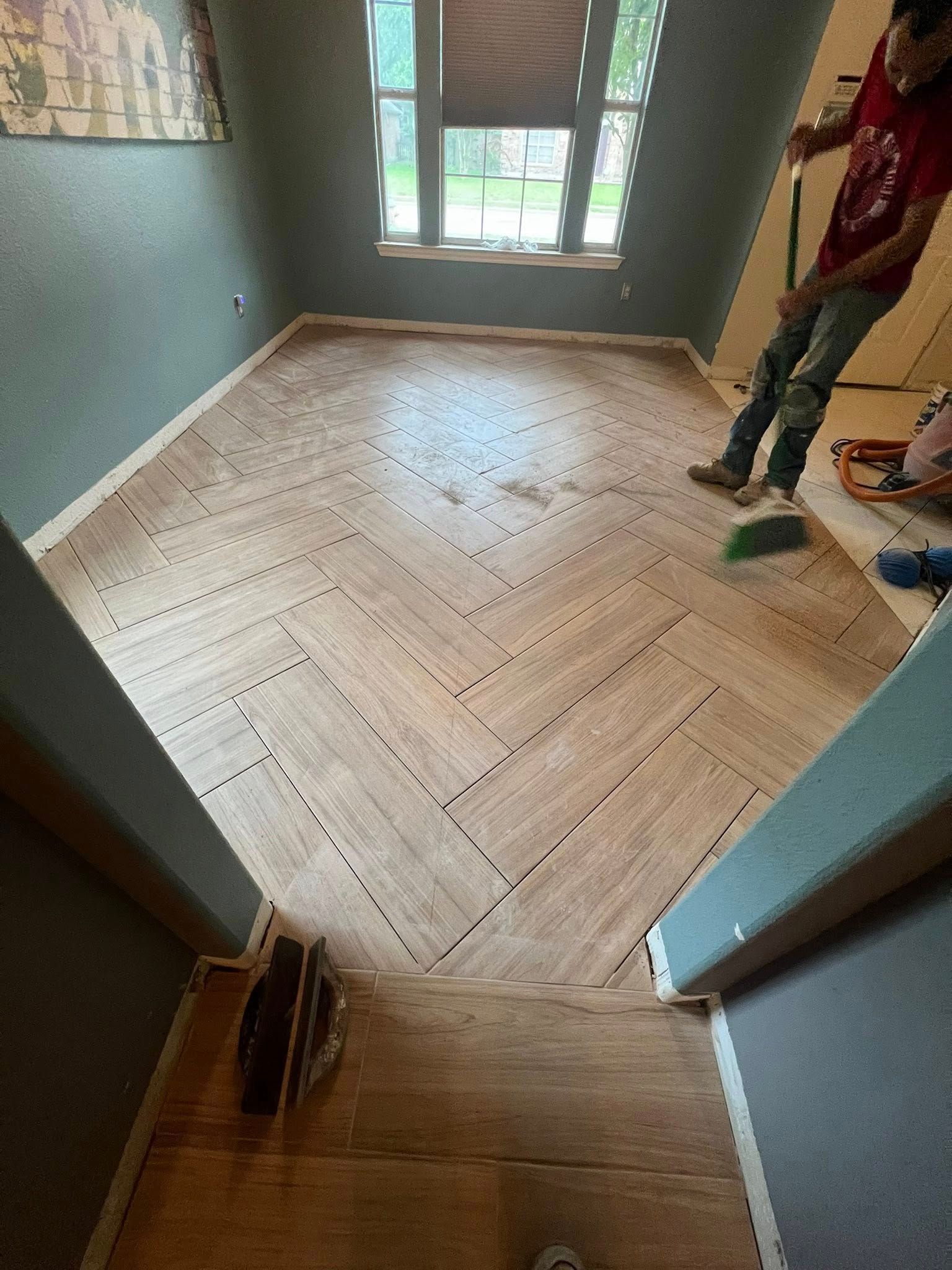 Herringbone wood floor installation in progress; worker sweeping; window in the background.