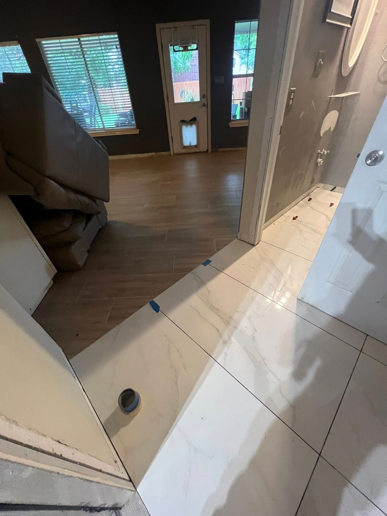 Looking from a tiled bathroom into a room with wood floors. A door and windows with blinds are visible.