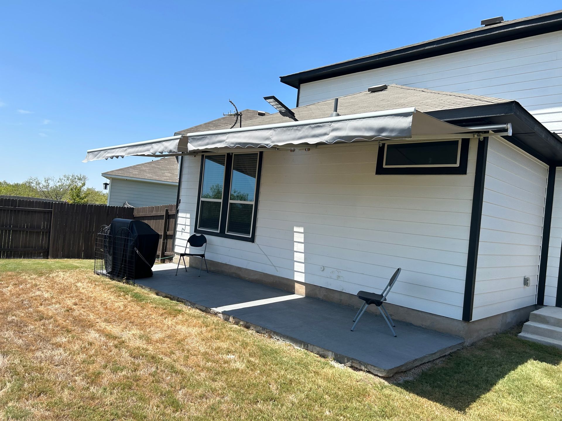 Backyard patio with retractable awning, white siding, grill, and chair. Sunny day.