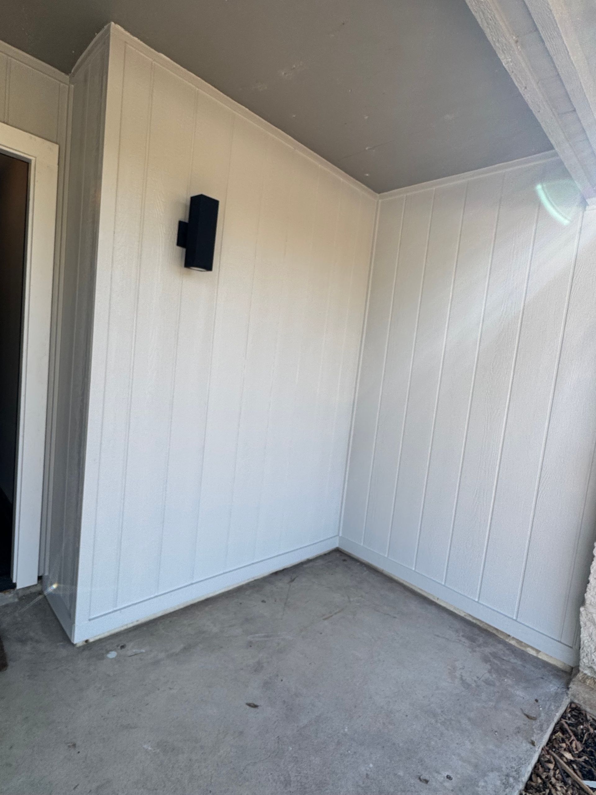 A small, partially-covered outdoor alcove with white paneling and a black light fixture on the wall. Gray concrete floor.