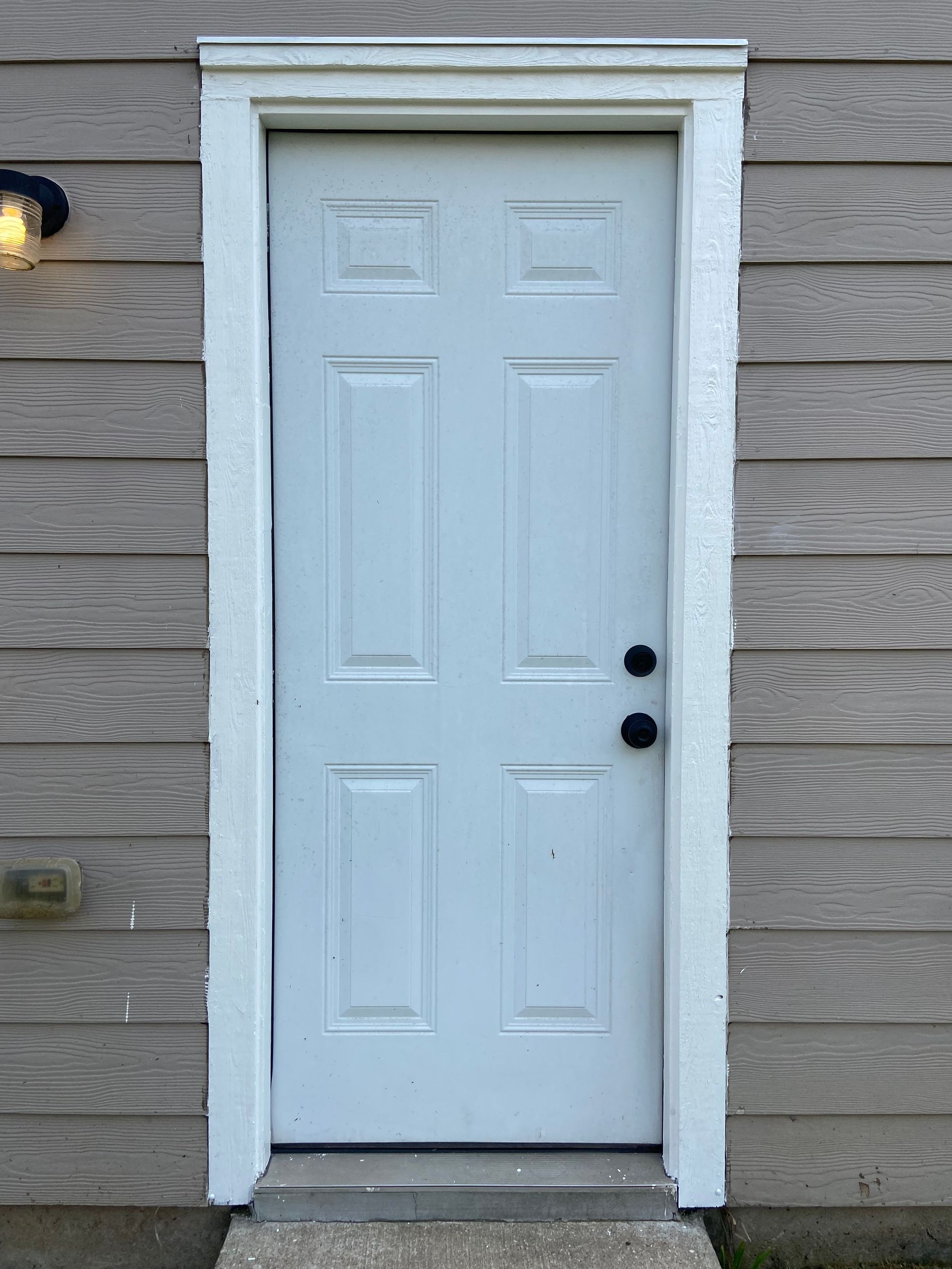 White six-panel door with white trim against a gray wood-paneled wall. Black doorknob, outdoor setting.