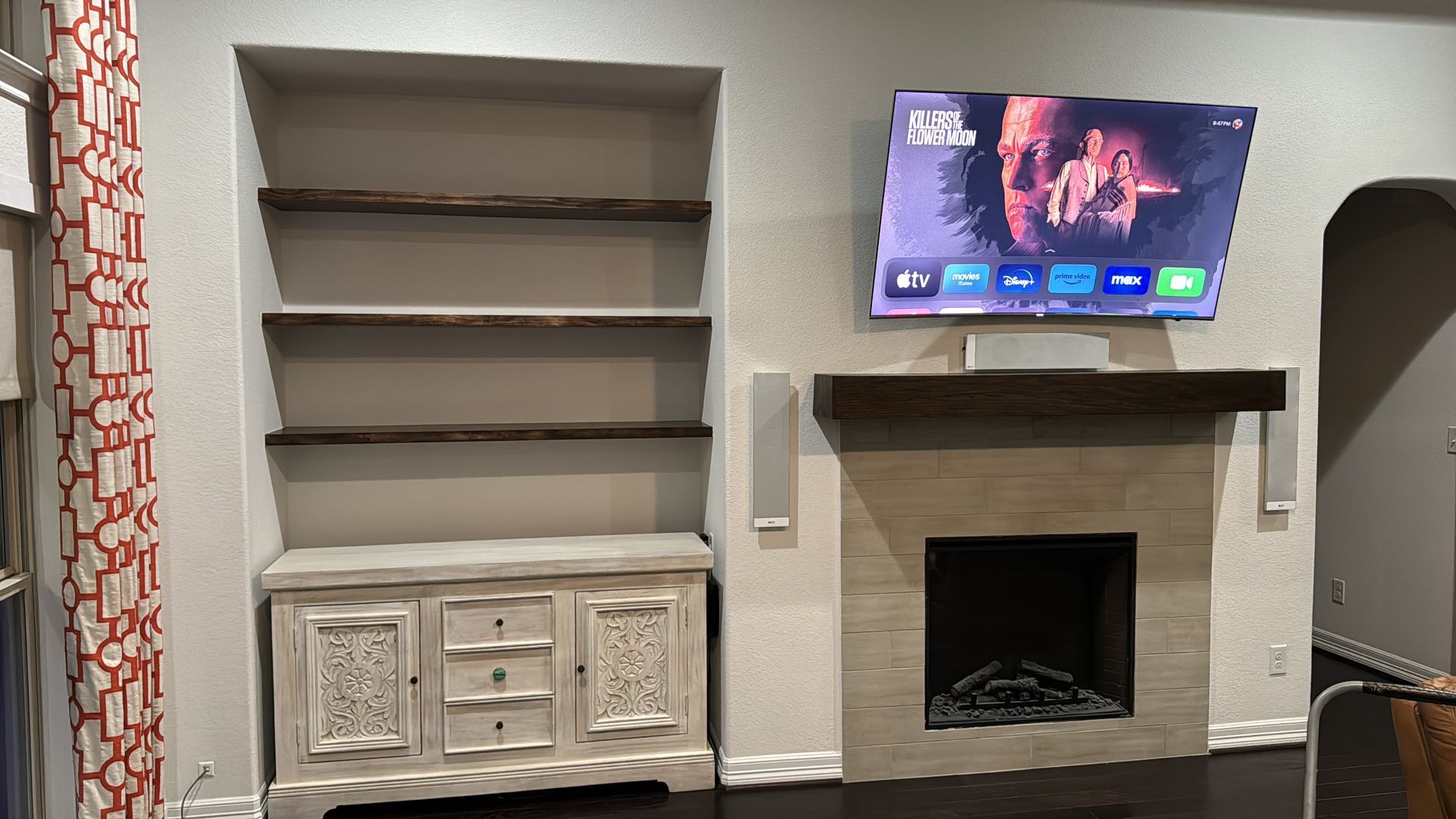 A living room with a fireplace & built-in shelving.A TV is mounted above the fireplace, with shelves & a cabinet to the left.