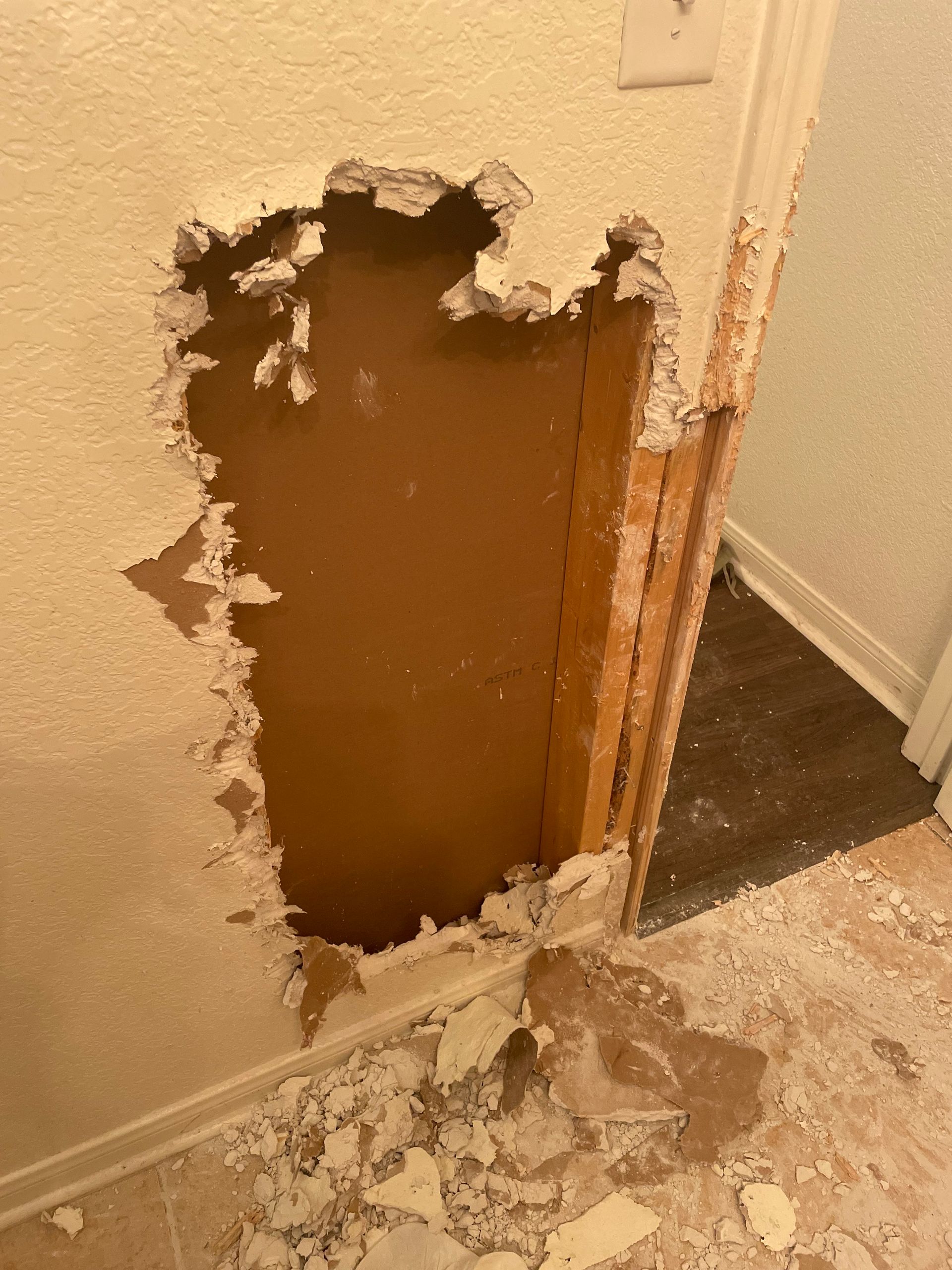 Hole in a beige wall exposing brown backing and wood studs, debris on the floor near a doorway.