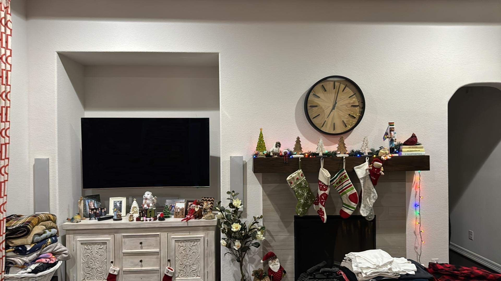A living room with a fireplace decorated for Christmas. A TV is above a white cabinet. A clock hangs above the mantel.