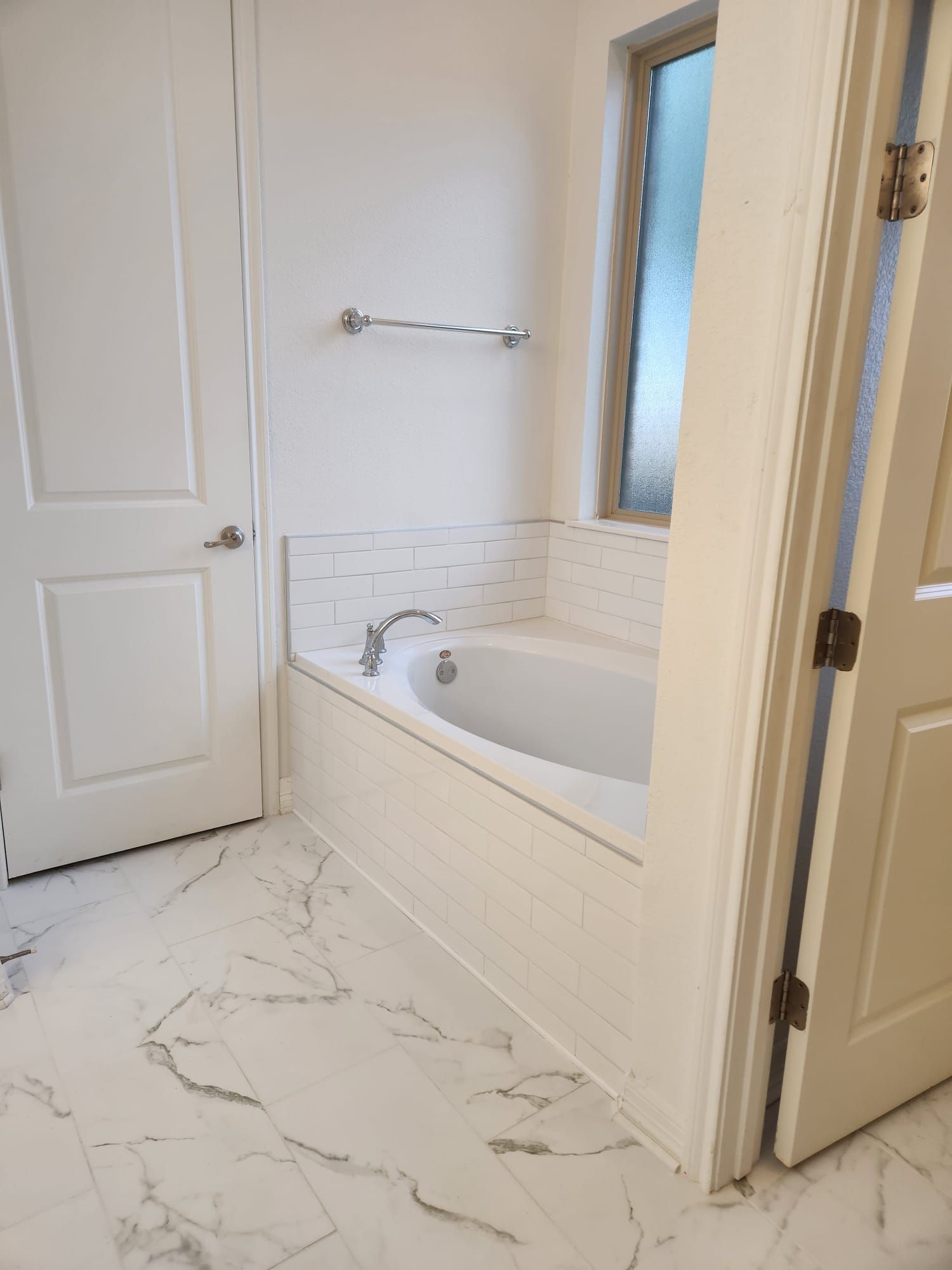 A bathroom with a white tub, white tile, and marble-patterned flooring. A closed door and a window are visible.