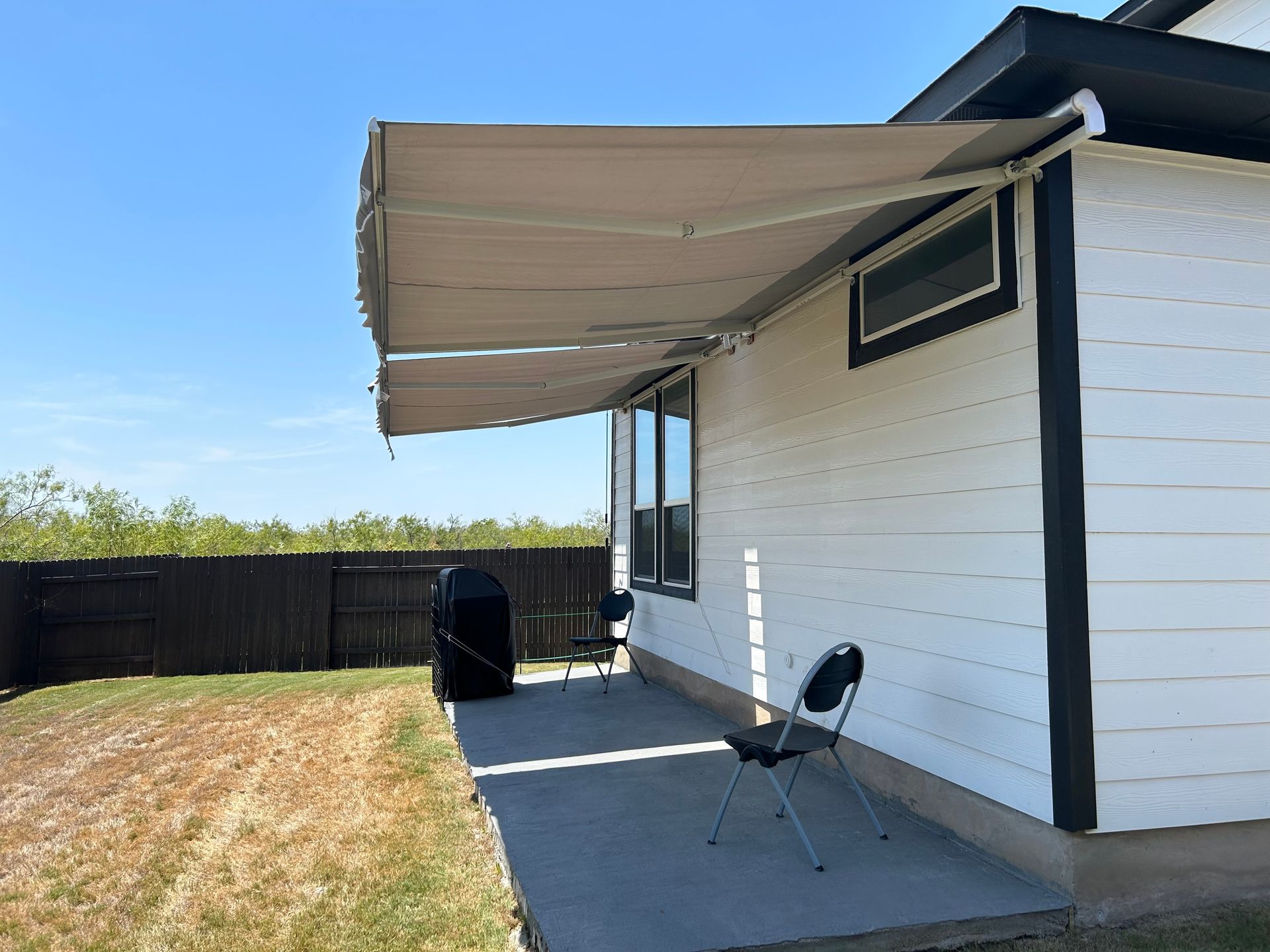 A beige retractable awning extends from a white house with black trim. A patio with a chair and grill sits below.