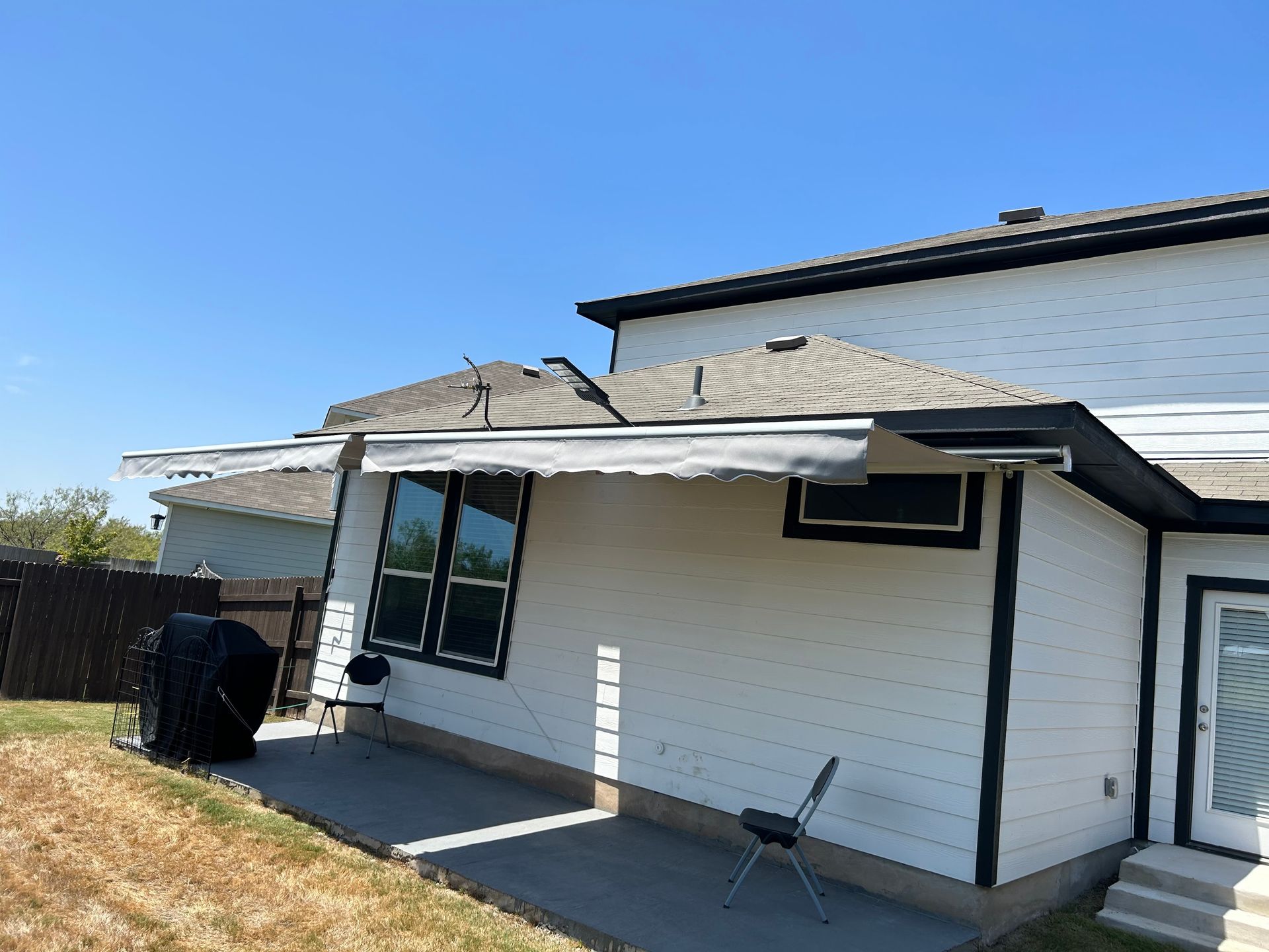 Backyard of a white house with a small patio, a grill, and a chair. The sky is blue.