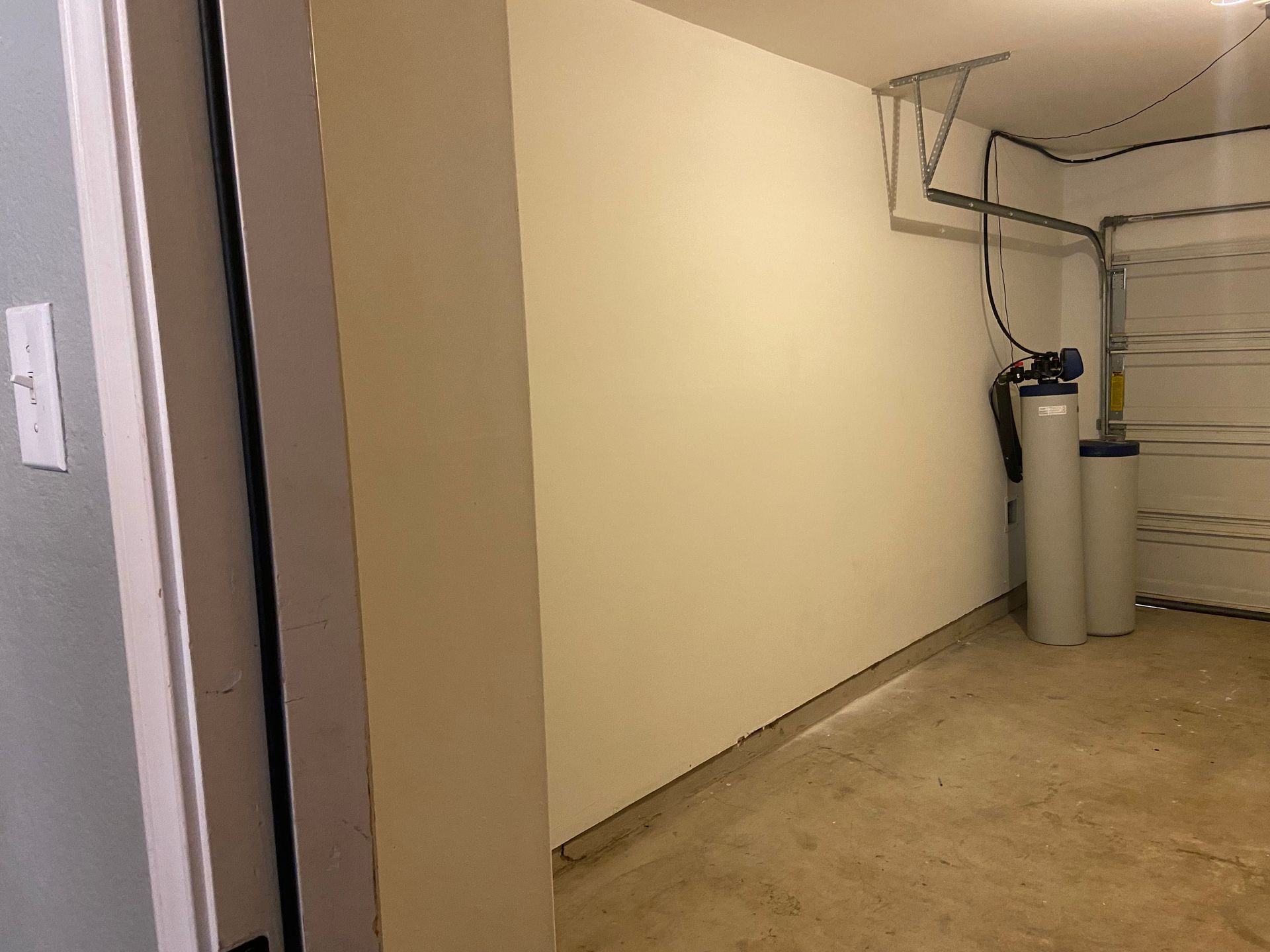 A light-colored garage interior. A water softener is visible against the back wall, and a door frame is on the left.