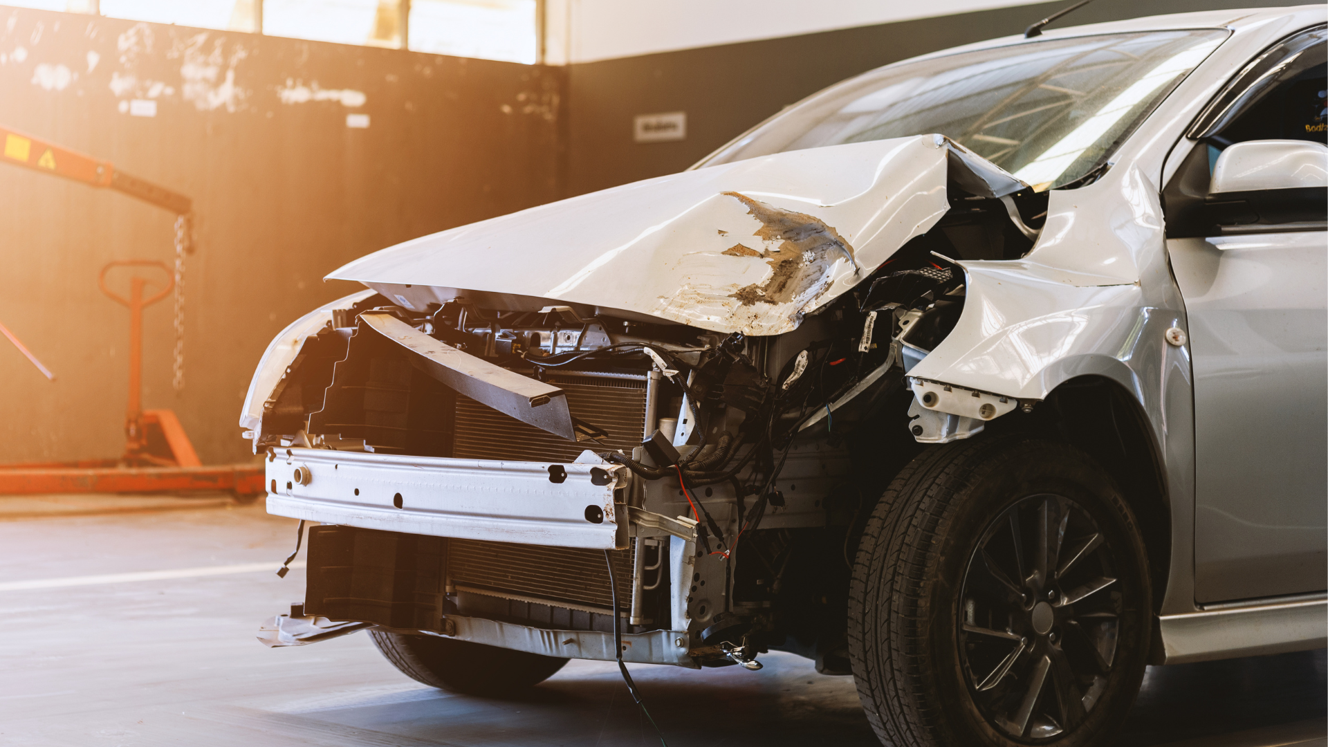 Damaged silver car, front end crumpled, likely after a collision, in an auto repair shop.