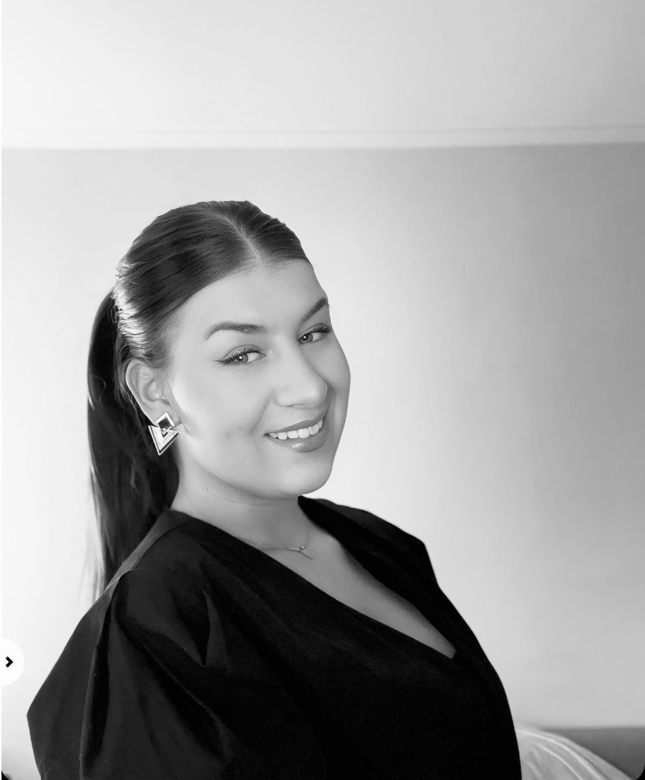 Een vrouw met donker haar in een paardenstaart lacht, ze draagt ​​een zwart shirt en oorbellen.