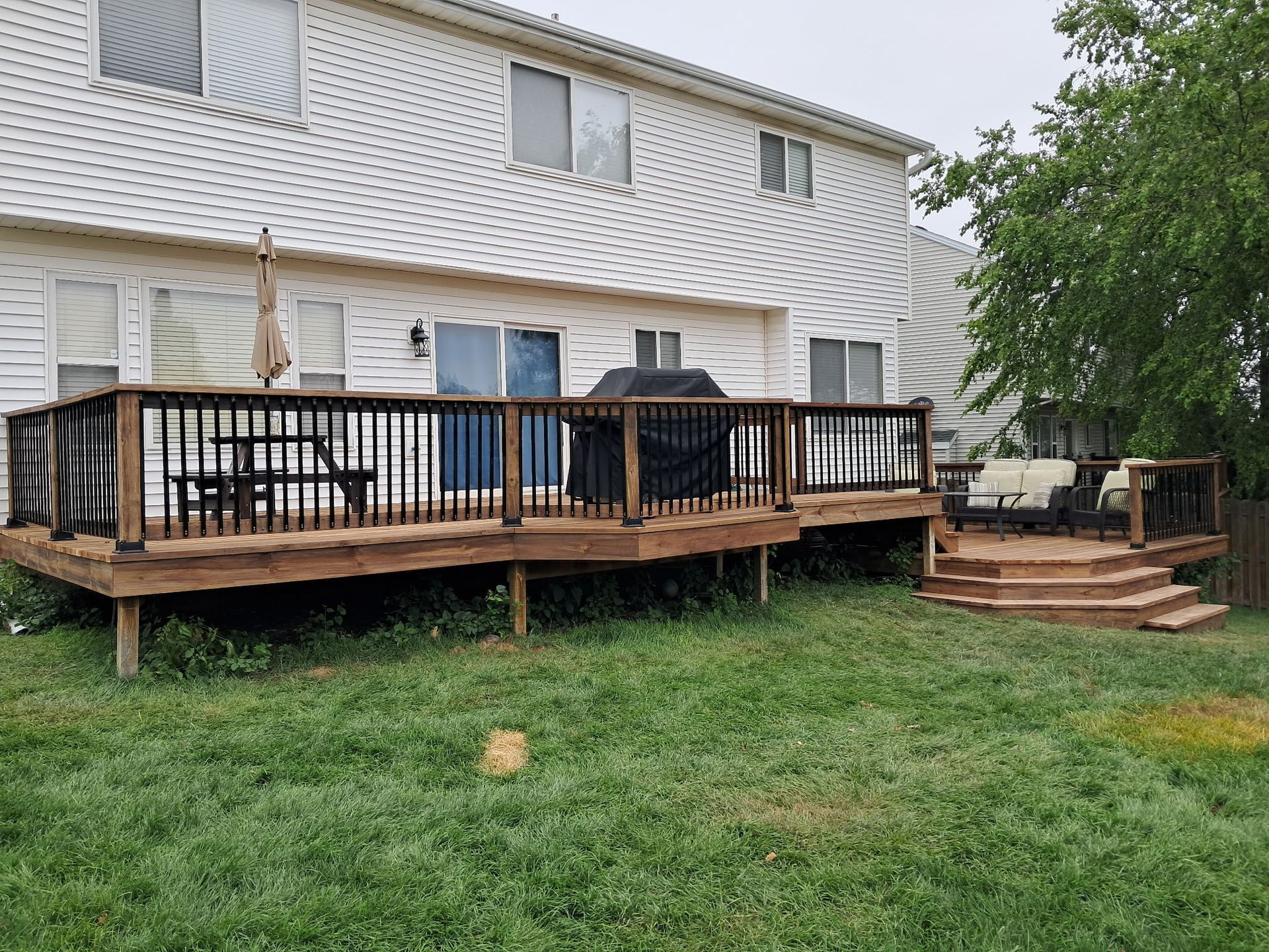 Backyard with multi-level wooden deck, black railings, grill, and outdoor furniture. Green grass and house in background.