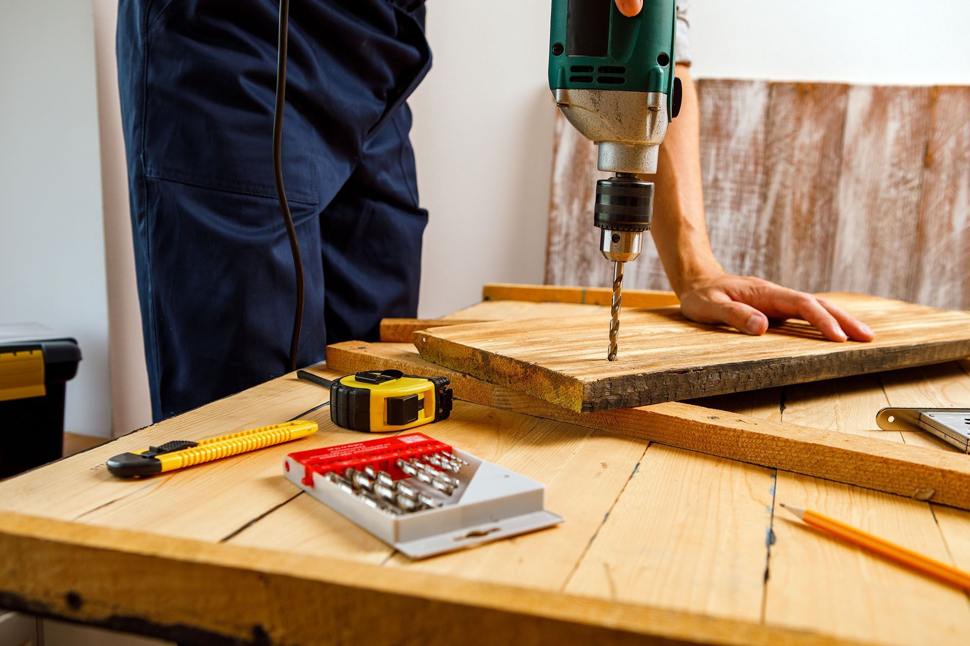 A person in navy work clothes uses a power drill on a wooden board, surrounded by tools on a workshop table.