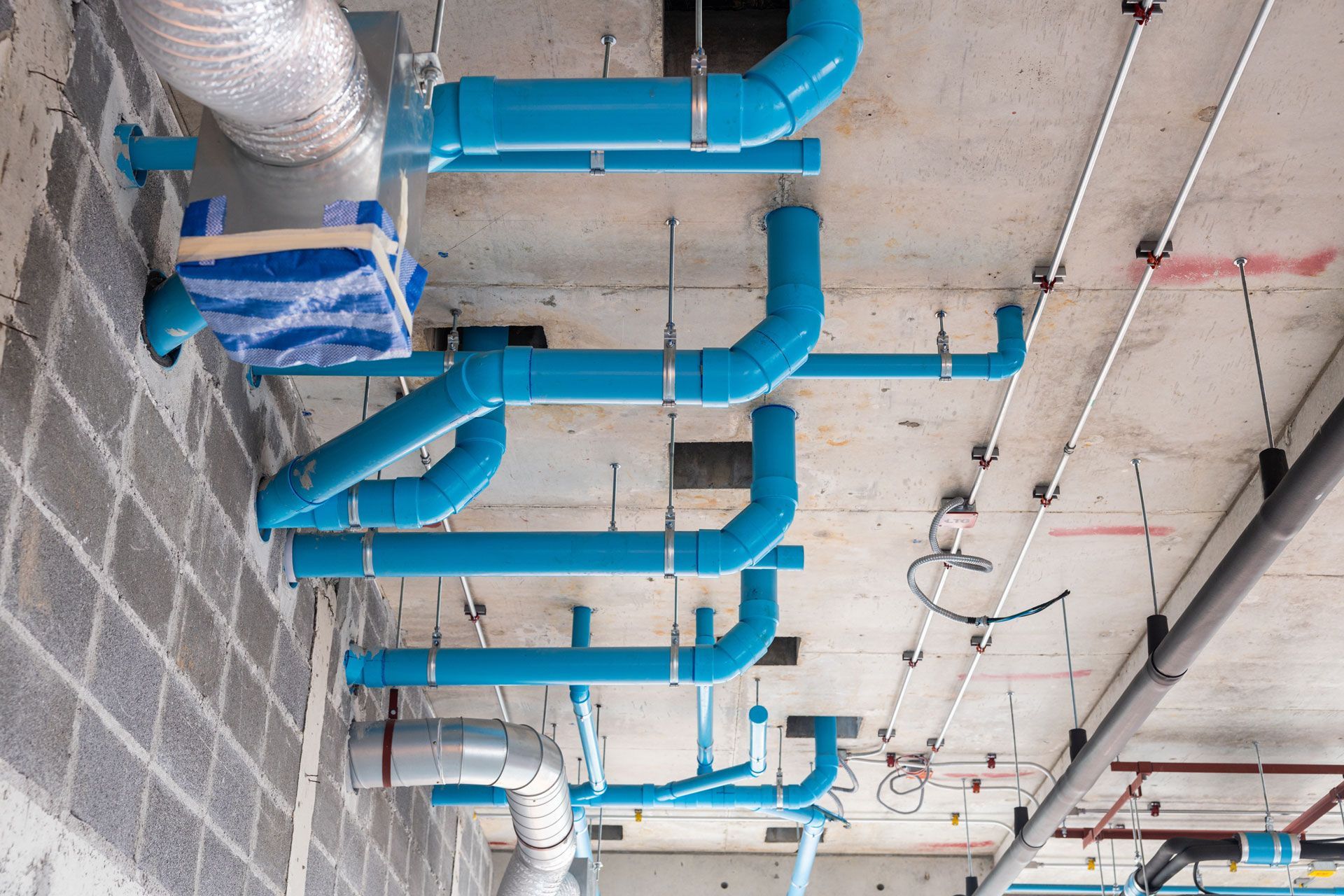 Blue pipes and silver ventilation ducts are installed along a concrete ceiling in an industrial building construction.