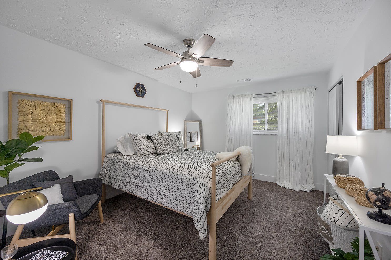 Bright bedroom in an apartment with a wooden bedframe, ceiling fan, and white curtains over a window.