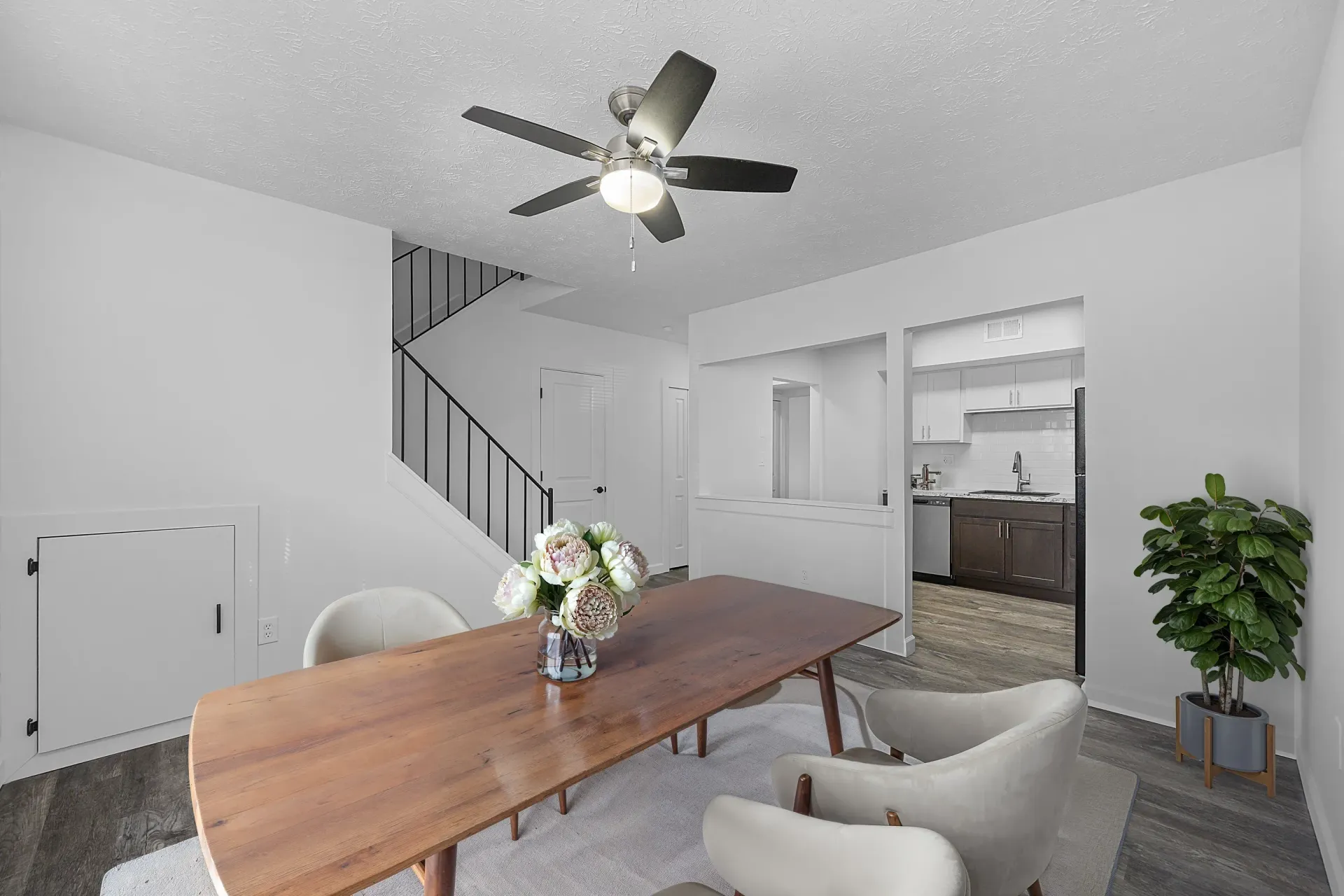 Dining area with wooden table, cream chairs, ceiling fan, and open kitchen with staircase nearby.