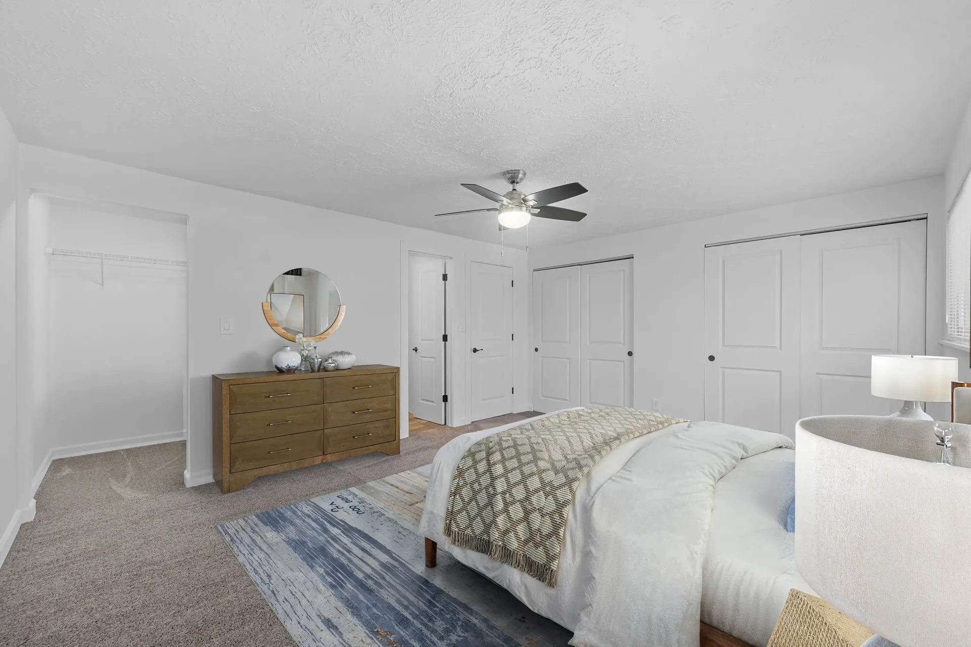 Bedroom in an apartment with a bed, dresser, round mirror, ceiling fan, and closet doors.