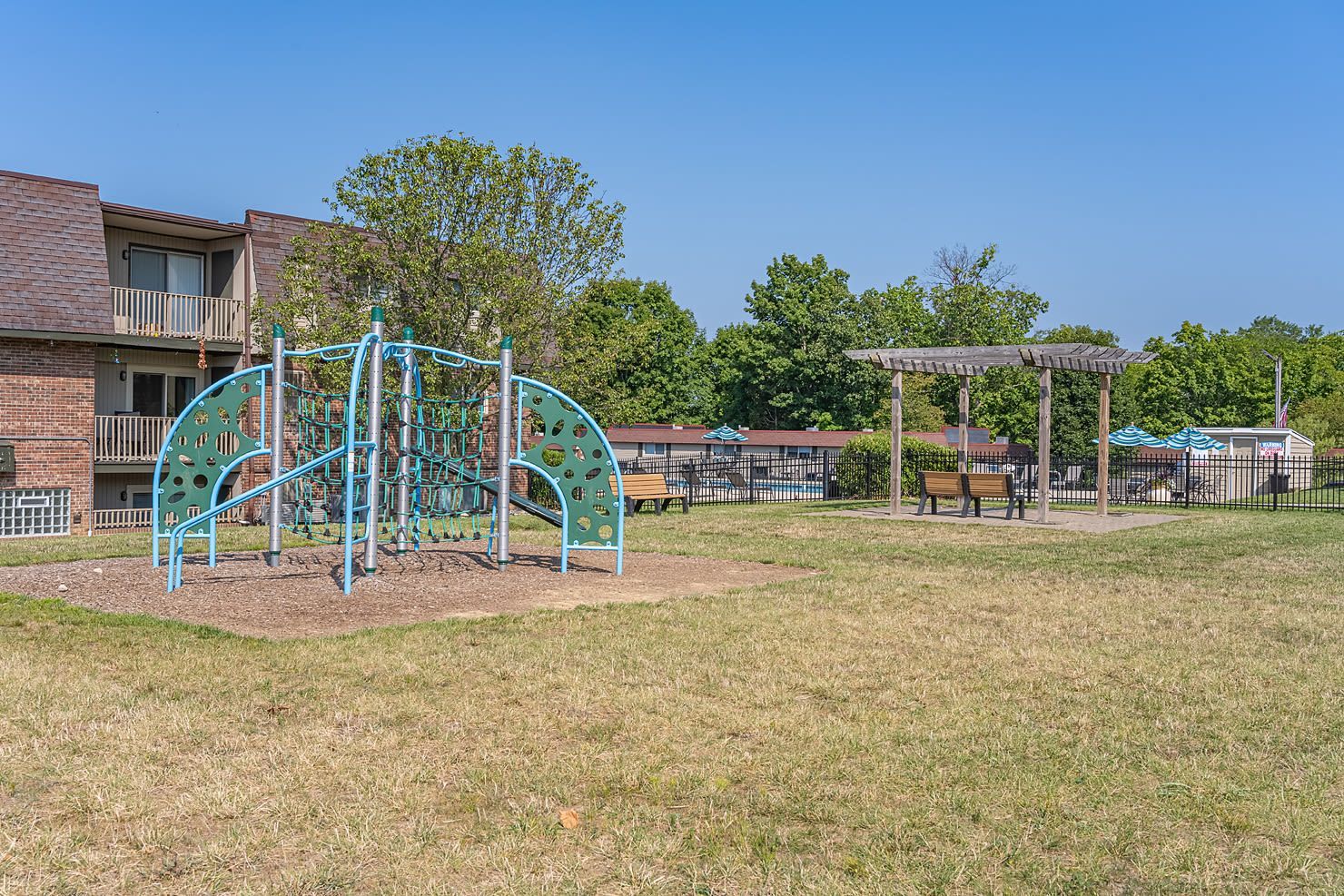 Playground structure in a grassy community yard with benches and a wooden pergola.
