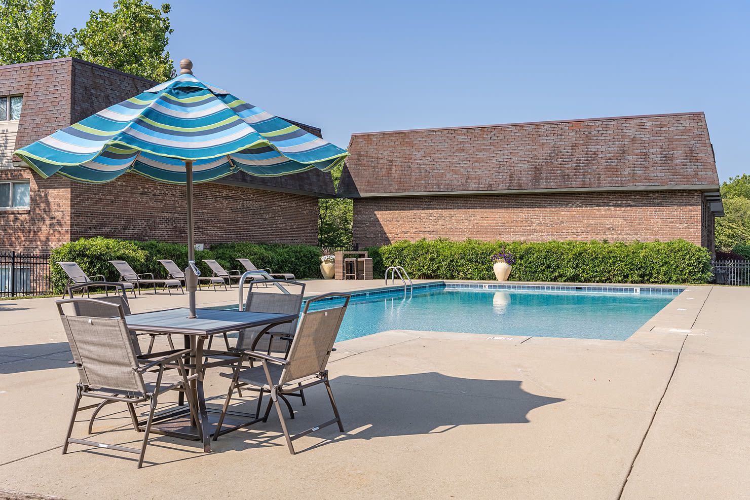 Outdoor apartment community pool with striped umbrella, lounge chairs, and brick buildings.